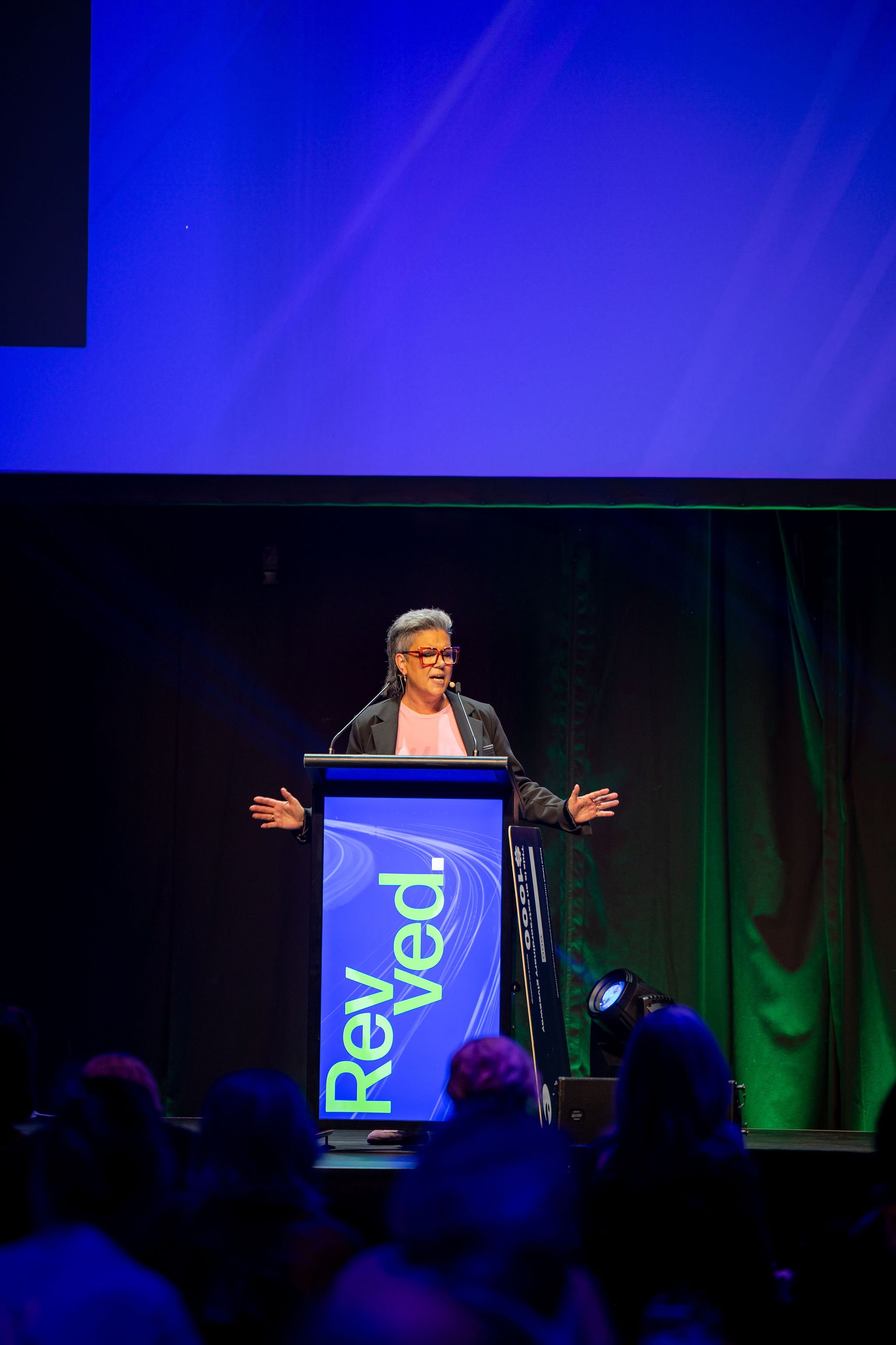 A woman standing at a podium giving a speech or presentation, with a large blue screen behind her and the event name partially visible on the front of the podium.