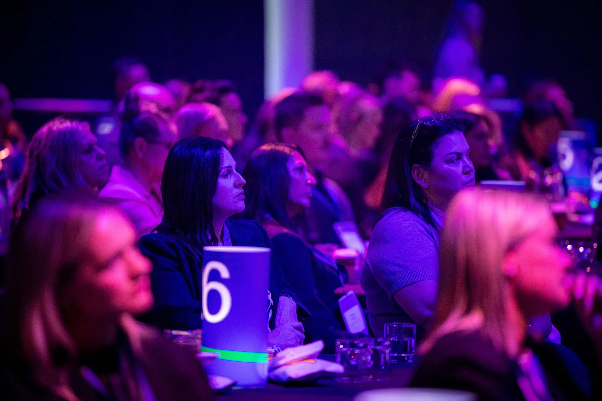 Audience members at Revved, focusing on a presentation or speaker, with purple and blue lighting.