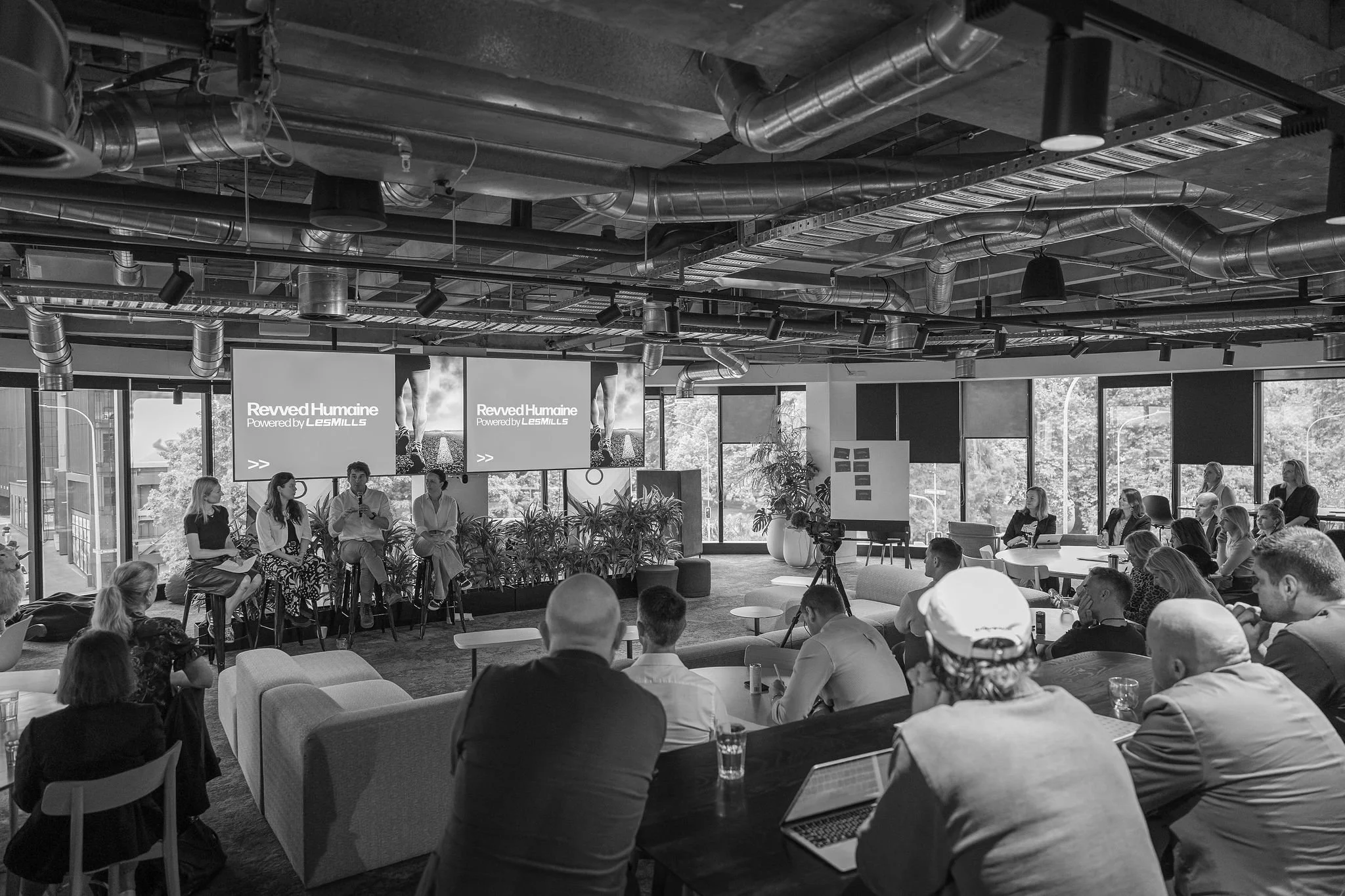A group of people attending a presentation or panel discussion in a modern conference room with large windows, multiple screens displaying 'Revved Humaine Powered by Les Mills,' and exposed ceiling pipes.