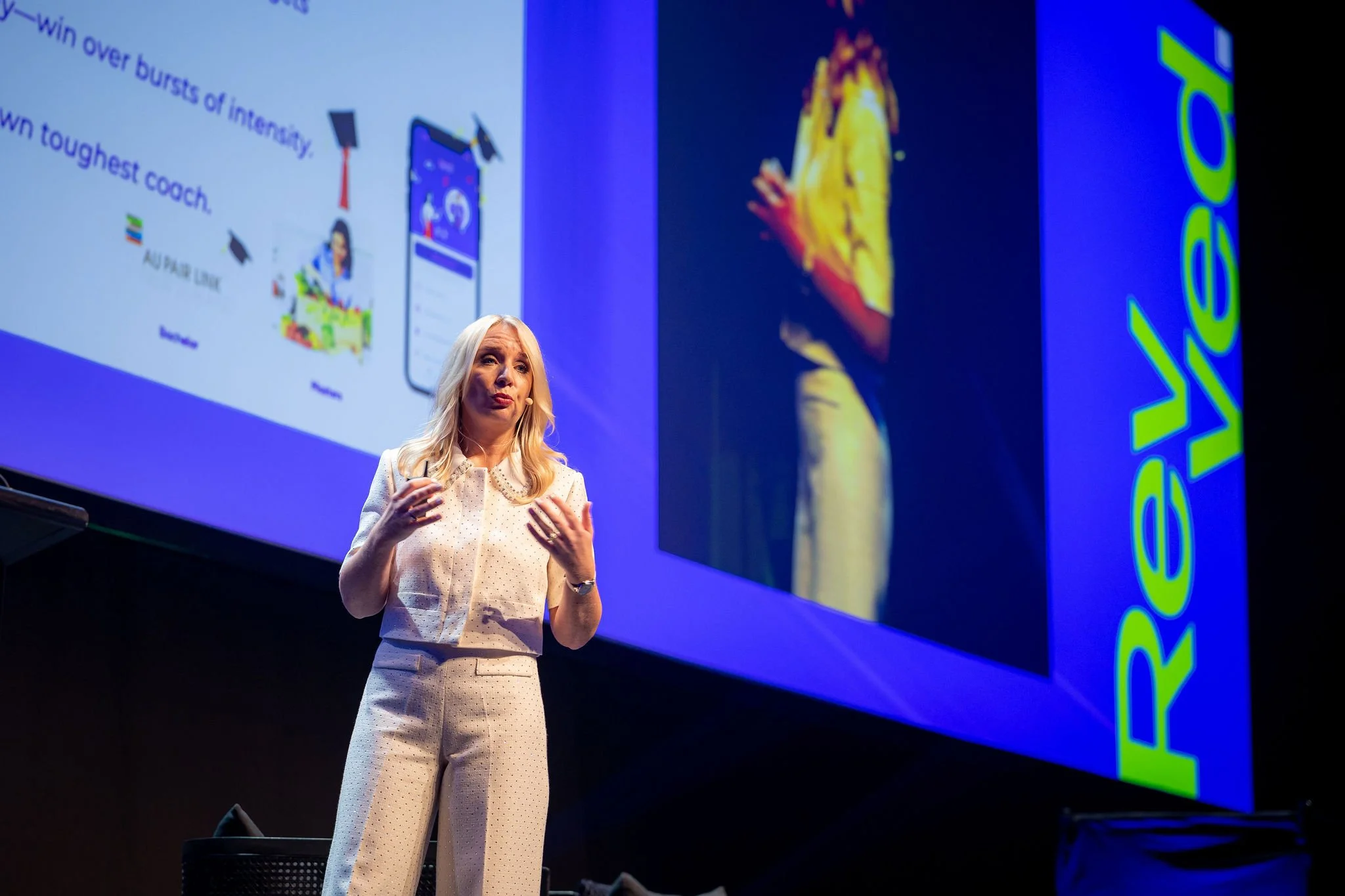 A presenter on stage at Revved, standing in front of a large screen displaying a colorful slide. The slide shows a mobile phone and a cartoon figurine with text. The woman is wearing a white dotted blazer and matching pants, gesturing with her hands.