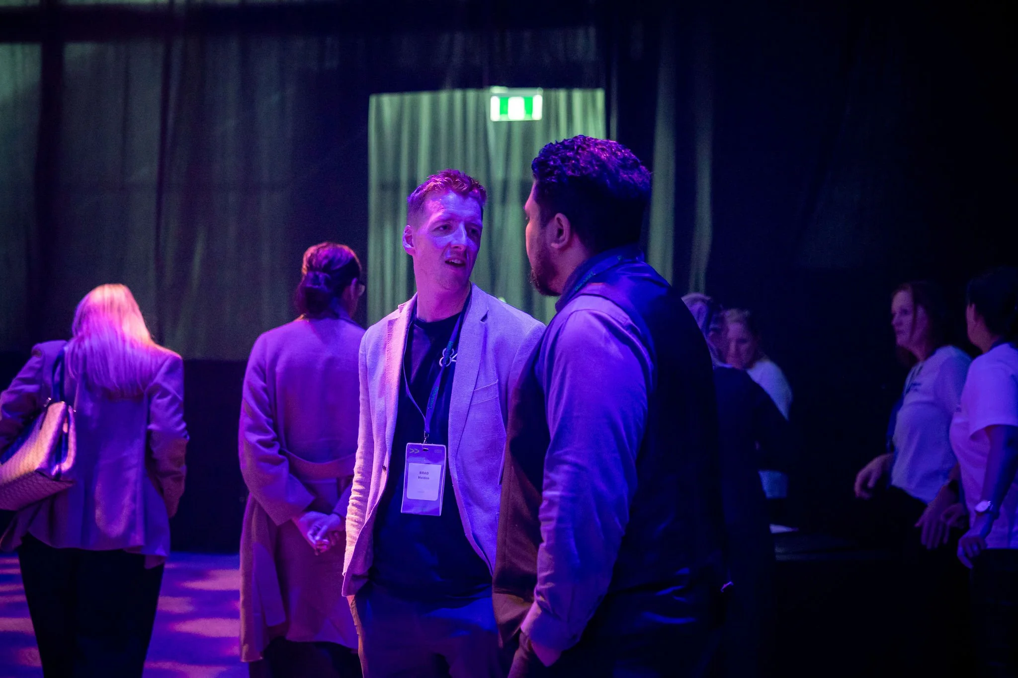 Two men are talking to each other at an event, surrounded by other attendees. The person on the left has red hair and is wearing a light blazer, while the person on the right has dark hair and a beard. They are engaging in conversation in a dimly lit