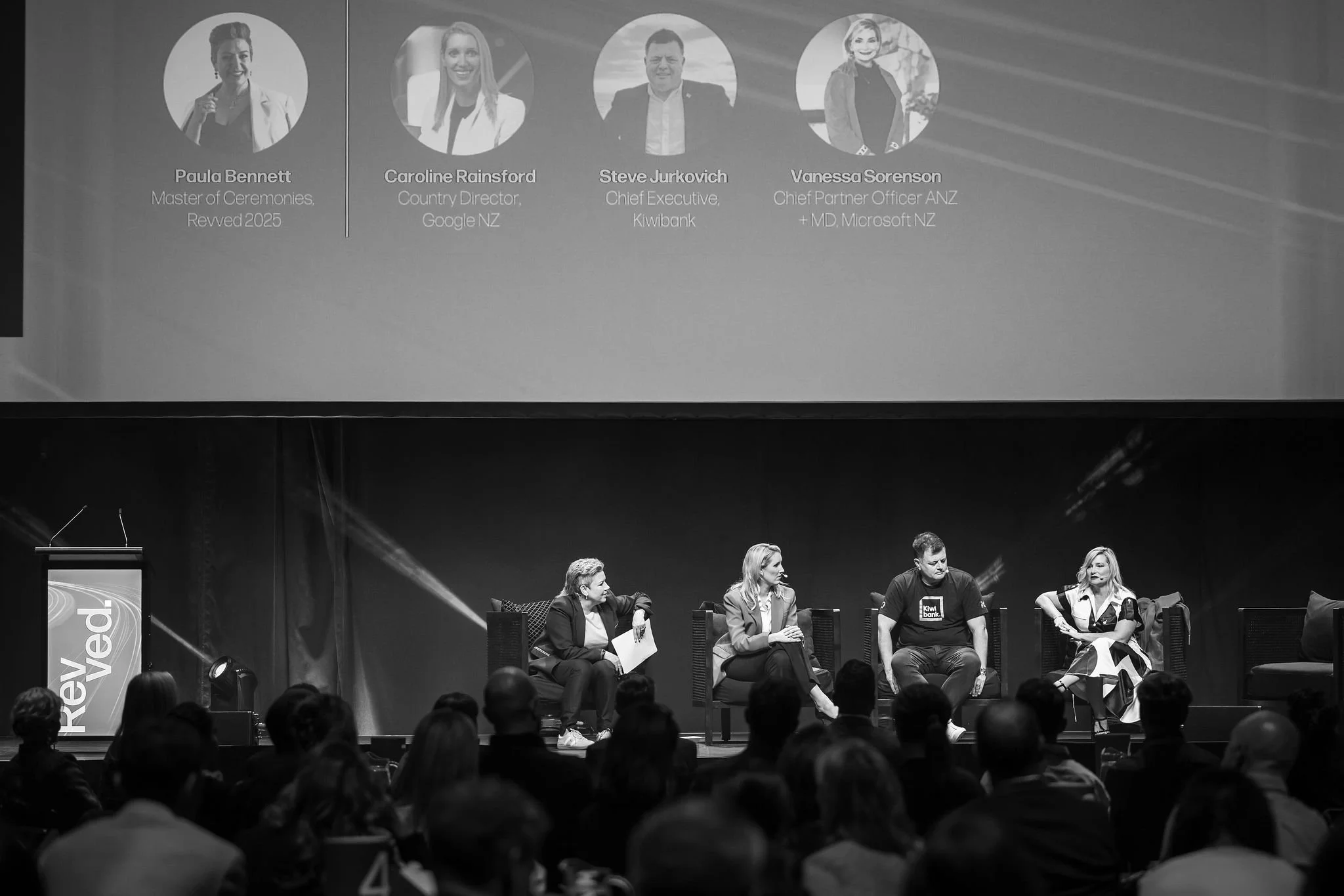 Black and white photo of a panel discussion on stage with five women seated in chairs, and a large screen overhead displaying headshots and names of four speakers, including Paula Bennett, Caroline Rainsford, Steve Jurkovich, and Vanessa Sorenson, wi