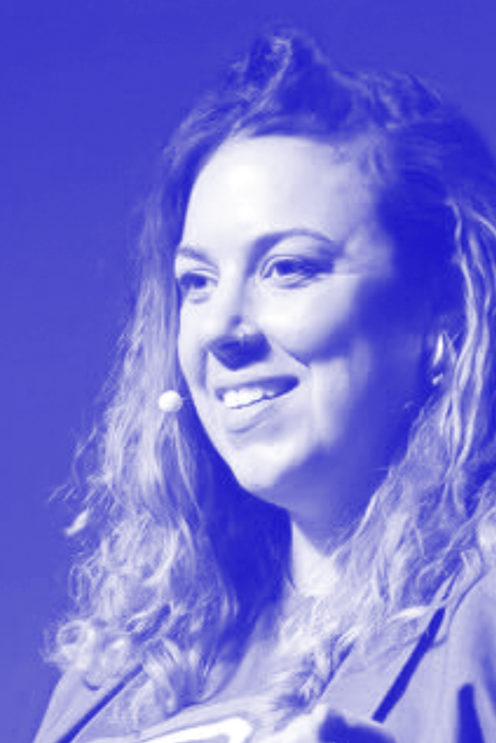 A woman with long, curly hair smiling and speaking on stage, wearing a blazer, with a dark background.