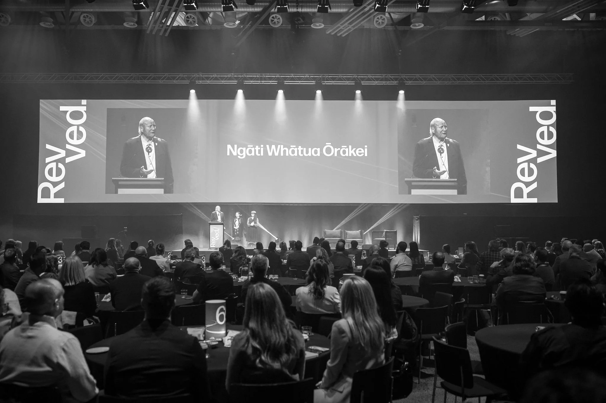 Black and White picture of the Revved stage with screens displaying the words "Revved" and "Ngāti Whātua Ōrākei." The audience is seated at round tables, some with name tags or numbers, and the venue has professional lighting and sound equipment.