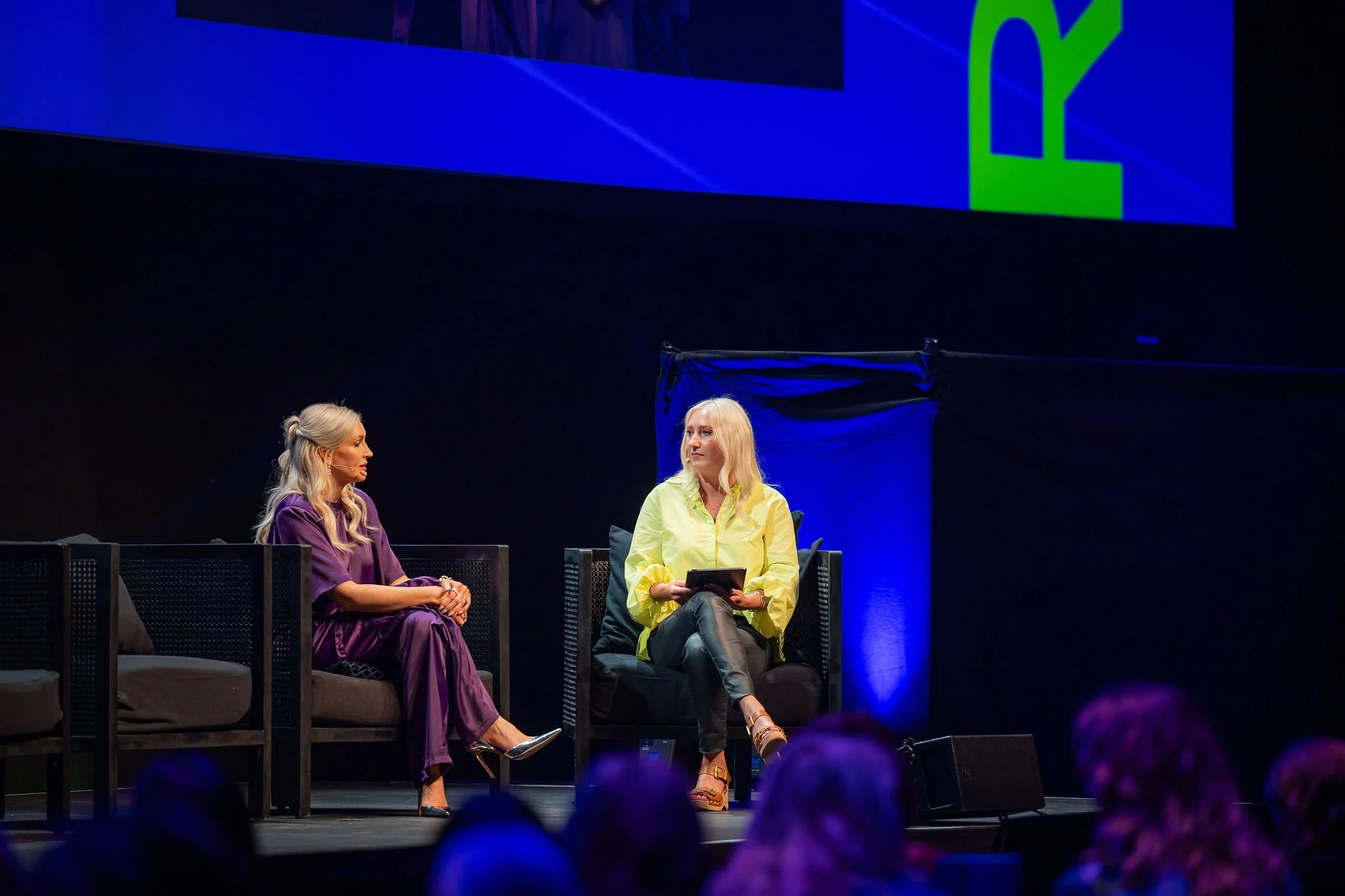 Two women sitting on stage at a panel discussion, one with long blonde hair in a purple outfit, the other with blonde hair in a yellow shirt holding a tablet, with a large screen behind them displaying a blue and green graphic.
