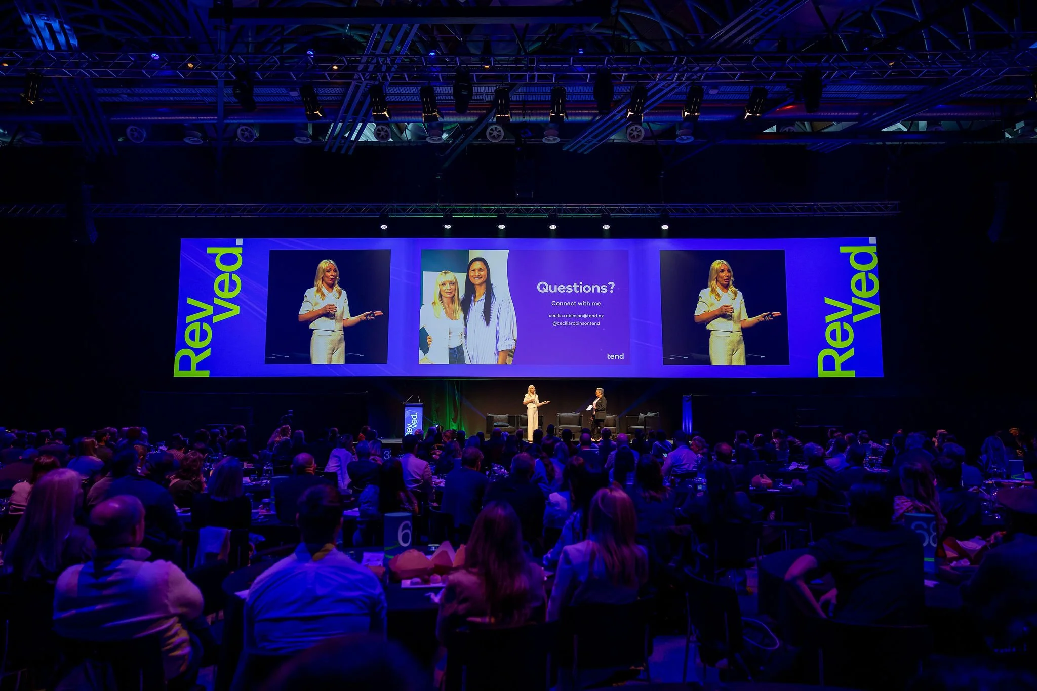 A large conference or seminar event with a stage featuring a large screen displaying two women and a question prompt. The stage has two presenters, a woman and a man, engaged in conversation. The audience is seated in rows, listening attentively. The
