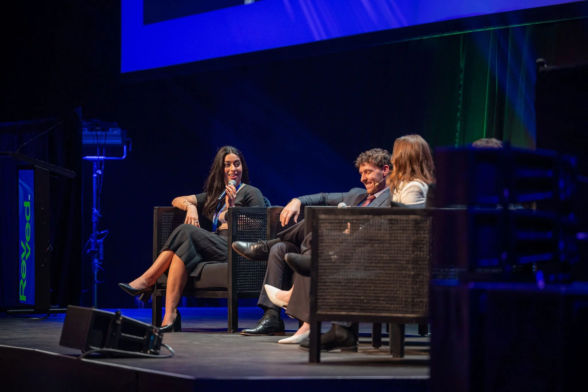 Three panelists on stage at a conference, seated and engaged in conversation. A woman with dark hair, black dress, and heels is speaking into a microphone, while a man with curly hair, suit, and tie and a woman with light brown hair and light-colored
