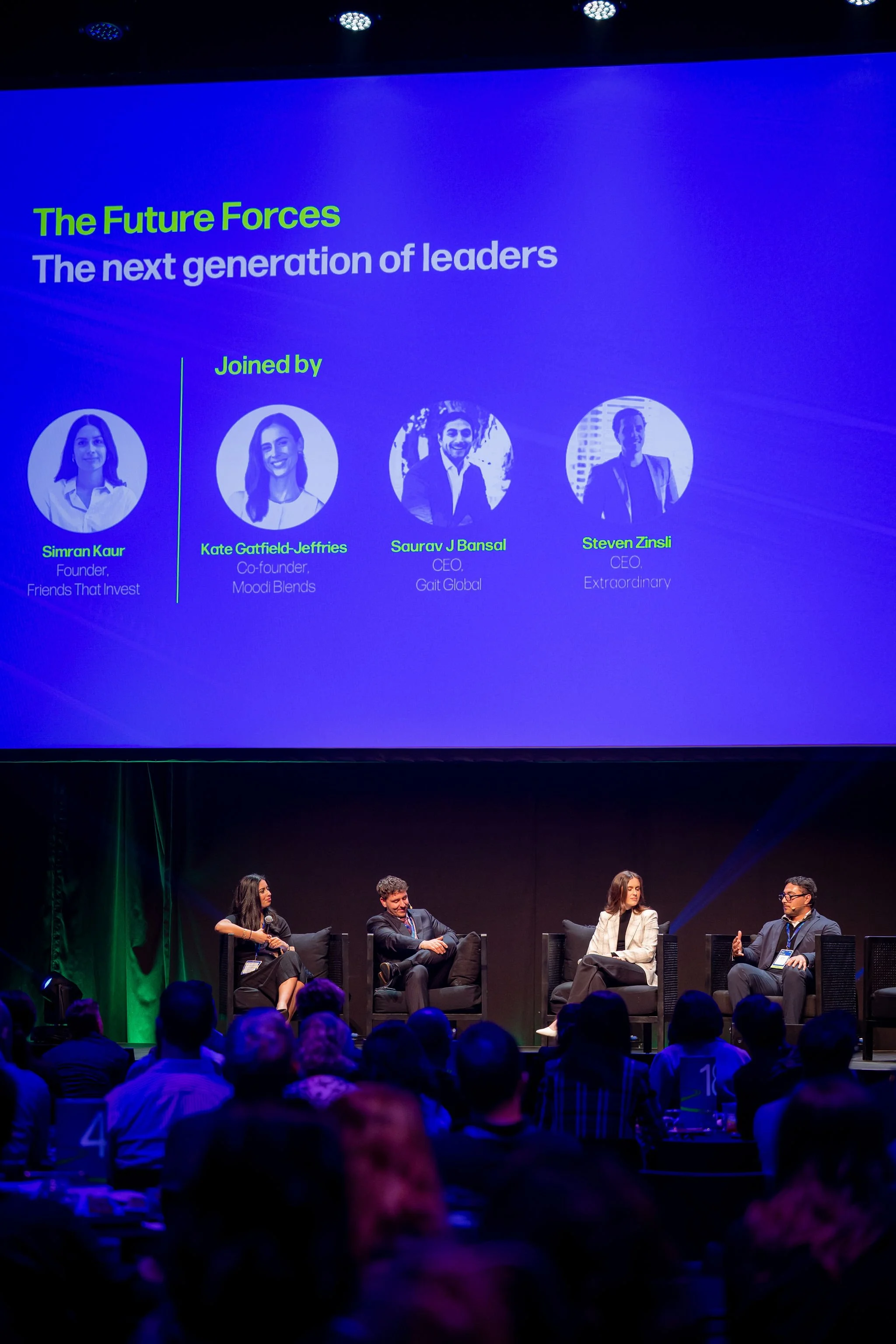 A panel discussion at a conference with four panelists sitting on stage and a large screen behind them displaying information about next-generation leaders, including their names and titles.