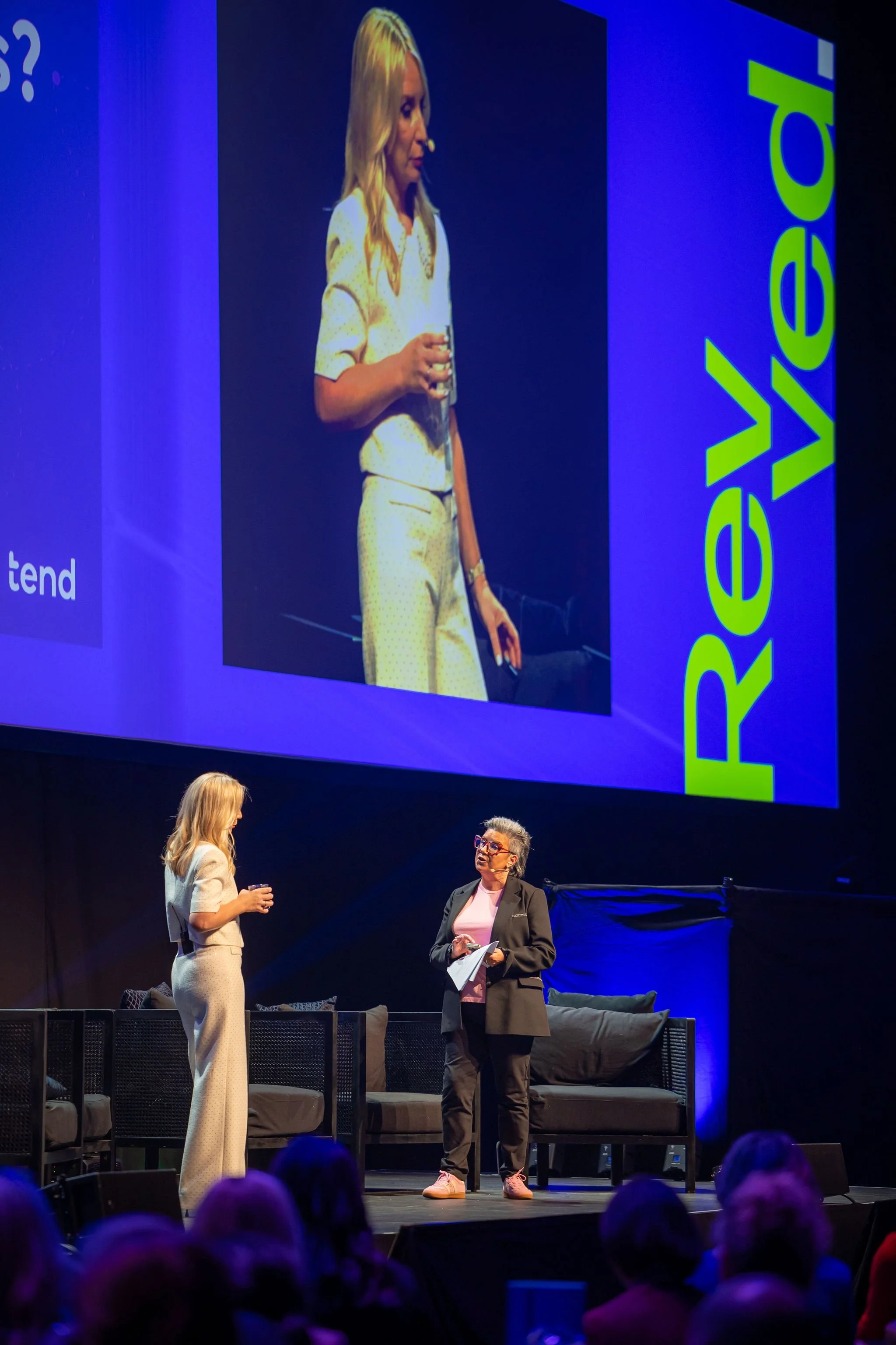 A speaker on stage with a microphone, facing another woman who is dressed in a dark blazer and pink shirt, while two large screens behind them display the same scene.