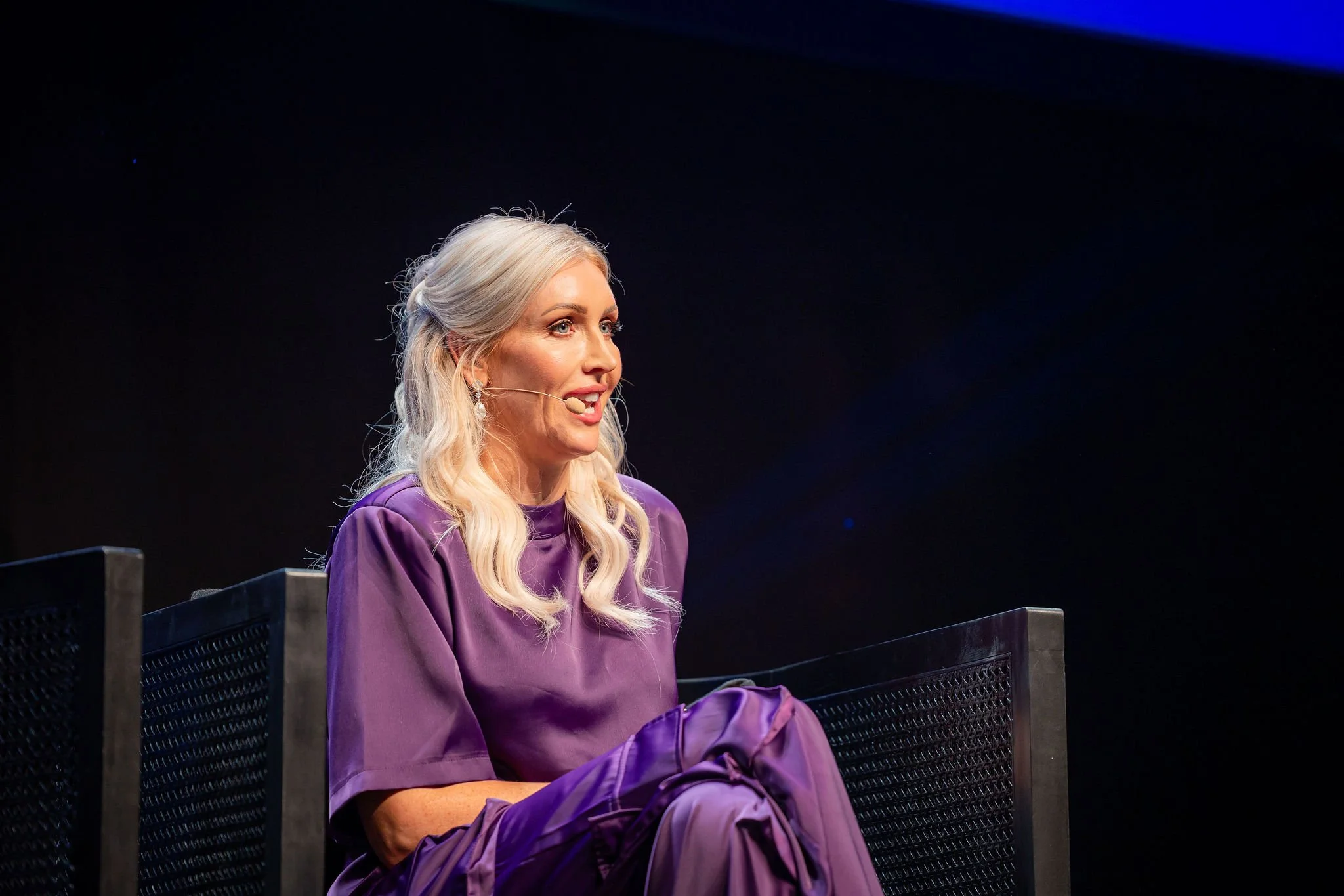A woman with long, wavy blonde hair, wearing a purple dress, speaking on a headset microphone. She is seated on a black chair with a dark background and blue lighting.