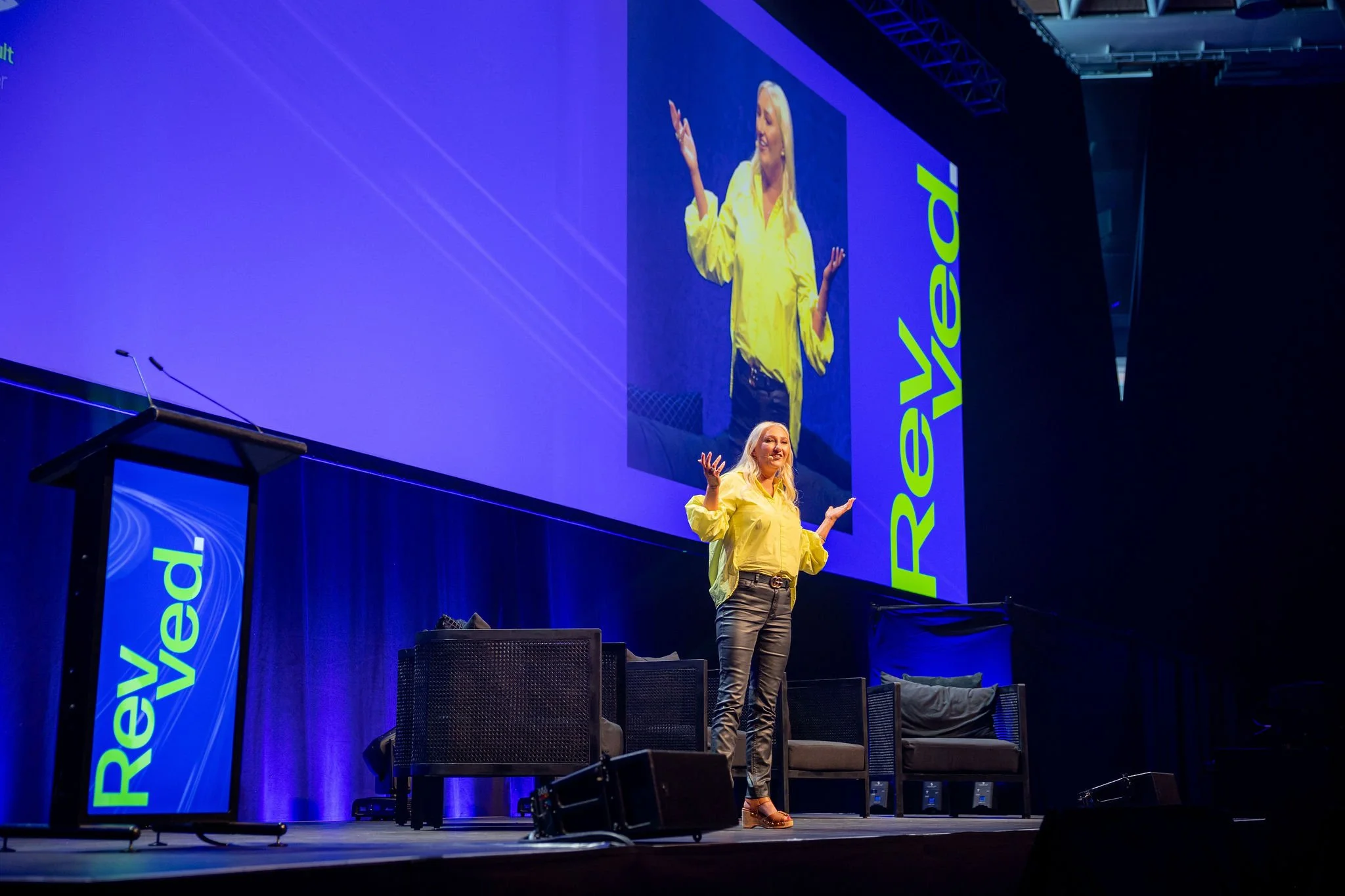 A woman with long blonde hair wearing a yellow blouse, dark pants, and high-heeled shoes is standing on a stage giving a presentation. She has her arms raised slightly and is smiling. Behind her, a large screen displays her enlarged image, and there 