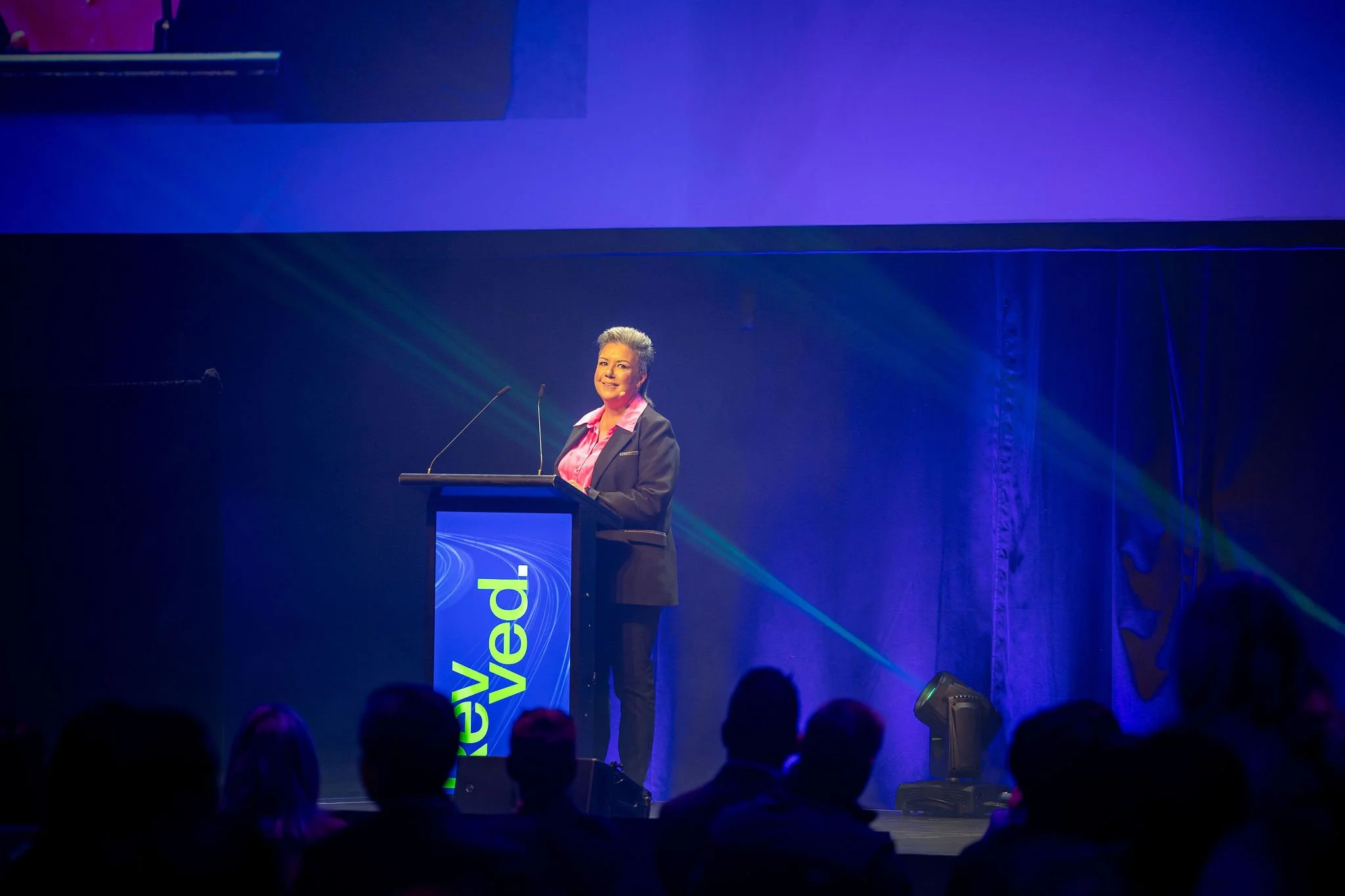 A woman with short gray hair wearing a suit and pink shirt speaks at the podium at the Revved conference., with a large dark backdrop and purple lighting.