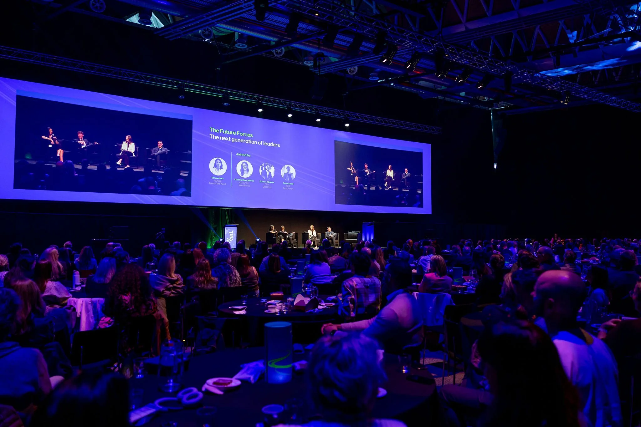 A large conference or seminar with an audience seated at tables, watching a stage with a panel of speakers and a large screen displaying a presentation titled 'The Future Forces: The next generation of leaders' with photos and names of four speakers.
