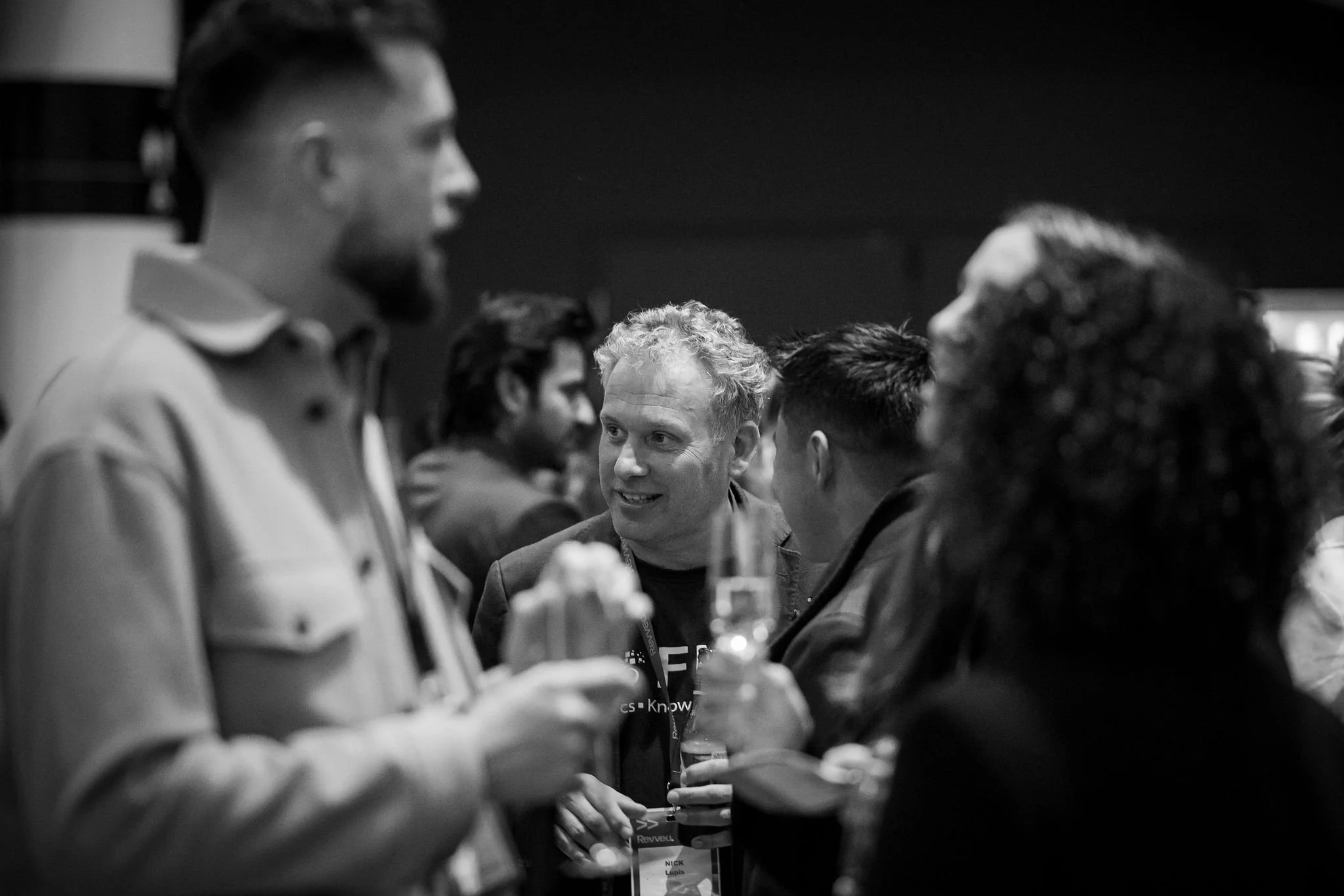 A black and white photo of a group of people at a social event, engaging in conversation and holding drinks.