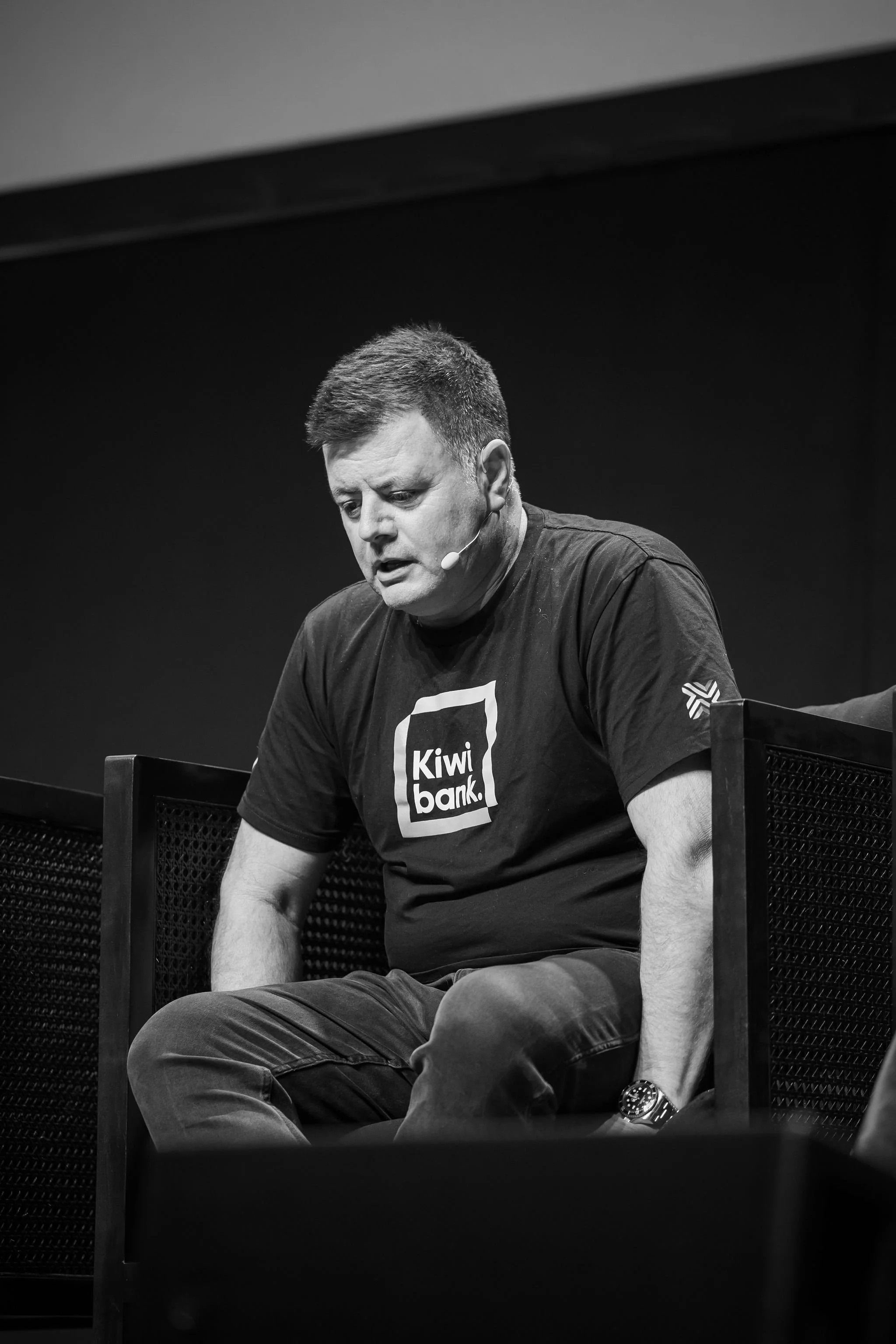 A man with short hair wearing a black T-shirt with the Kiwi Bank logo, sitting on a chair, looking down, with a microphone headset, in a dark room.
