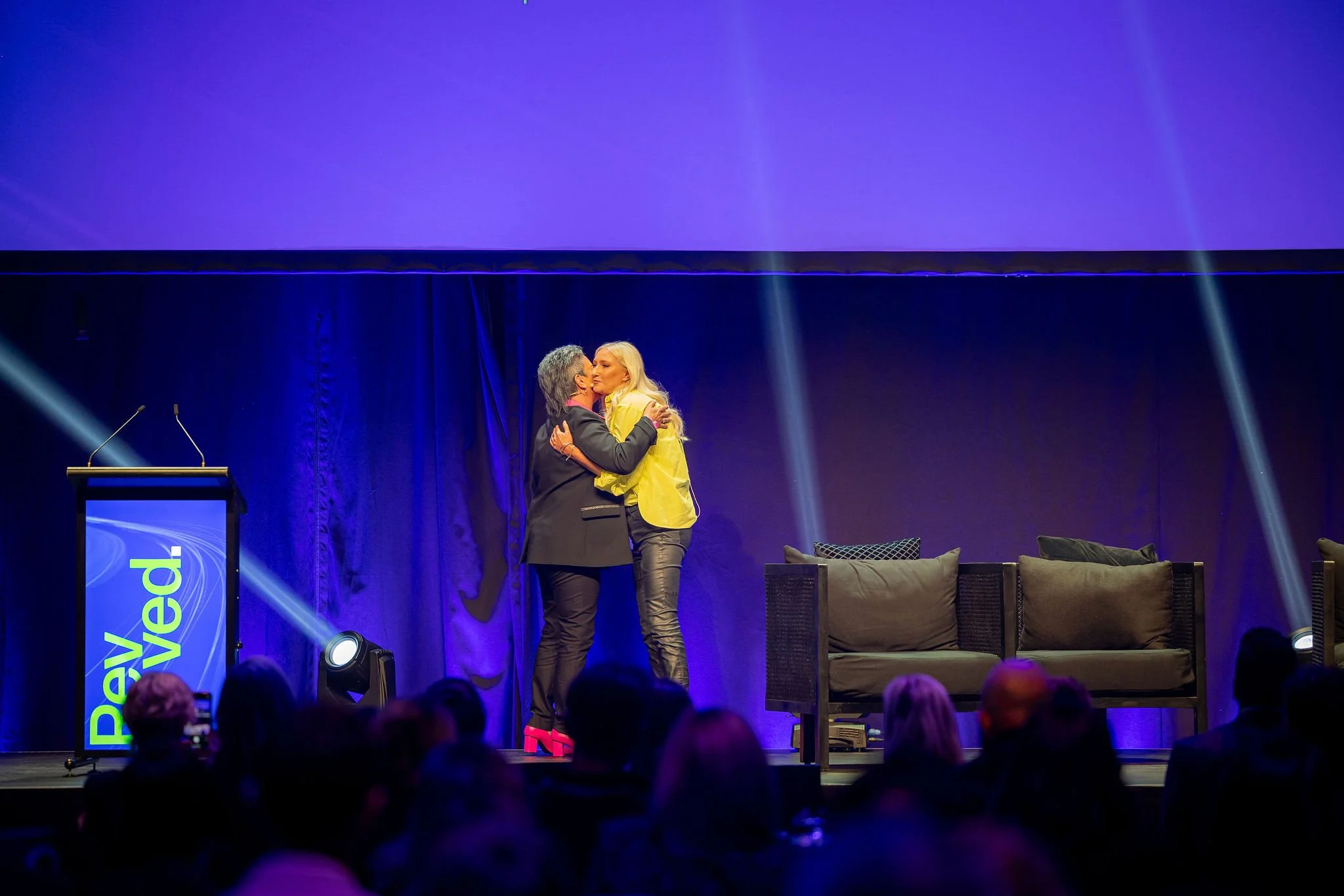 Presenters on stage at Revved. The stage has purple and blue lighting with a seating area, and an audience is visible in the foreground.