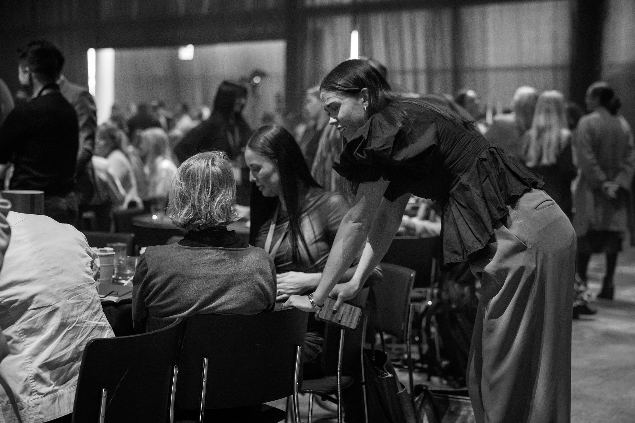 A woman is leaning over a table, engaging in conversation with a child with shoulder-length hair in a busy conference or event setting with many people seated and standing around.