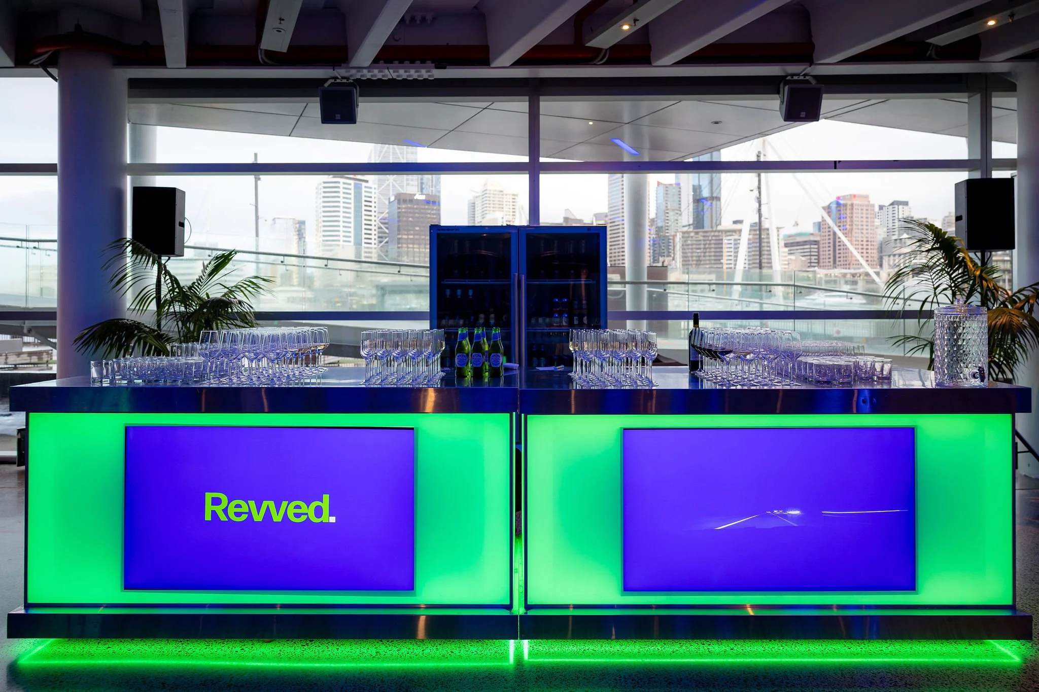 A modern bar counter with a bright green illuminated front and a large digital display on it. The display shows part of the text 'Revd.' in green and blue colors. Behind the bar, there are empty glasses and bottles, with city buildings visible throug