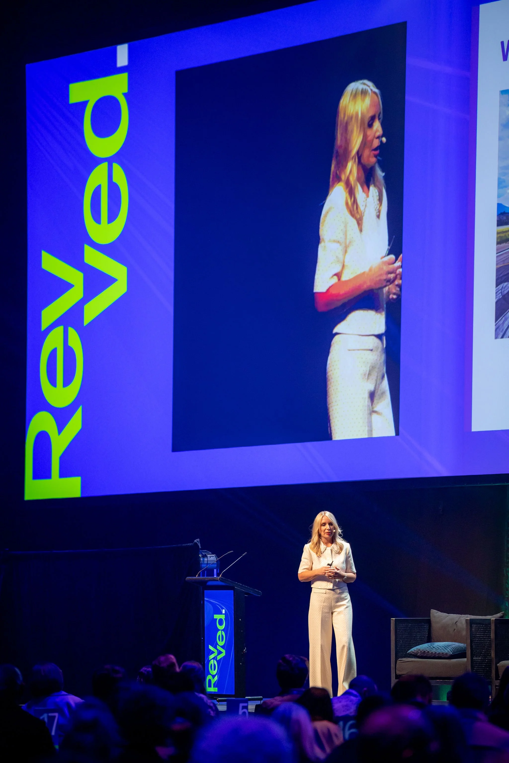 A woman in a beige suit is speaking on stage at a conference or event, with a large screen behind her displaying her image and a vertical blue sign with the word 'Revealed' in green letters.