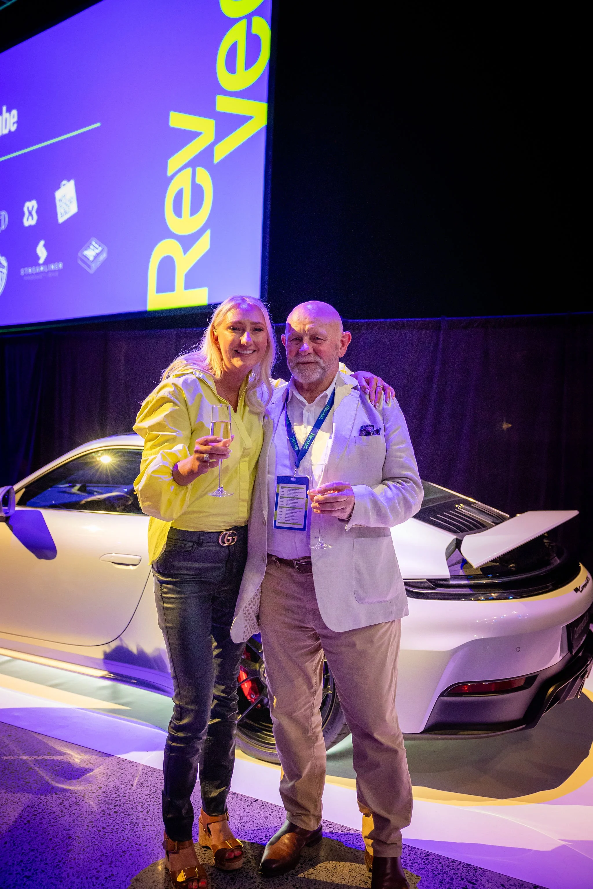 A woman and an older man stand in front of a white sports car at a corporate or industry event, smiling and holding glasses of champagne. Behind them, a large screen displays the logo and branding of REVE, with various icons. The setting is dimly lit