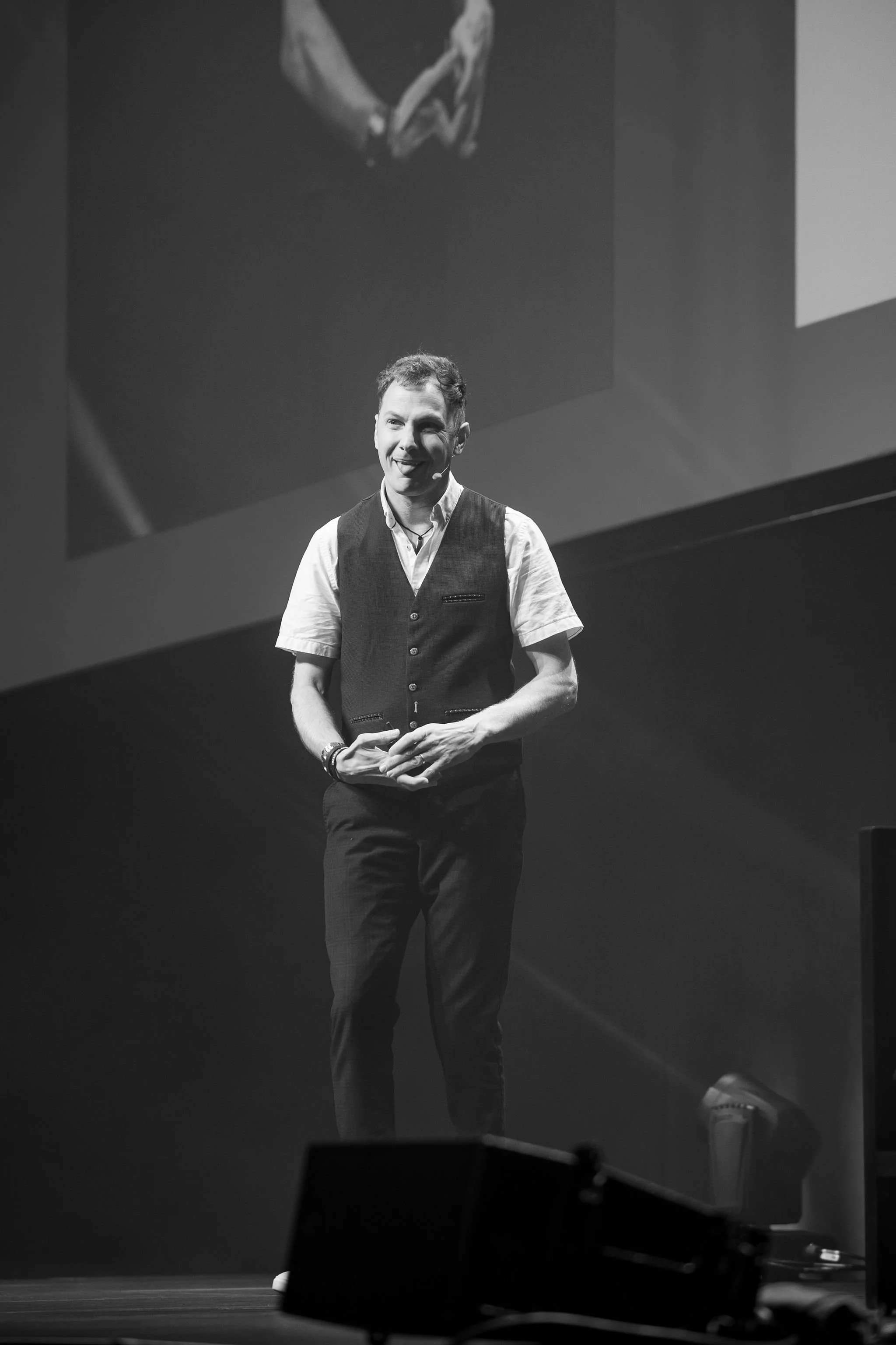 A speaker on stage at Revved, smiling and standing in front of a large screen showing his live image, dressed in a vest and shirt, with stage equipment in the foreground.