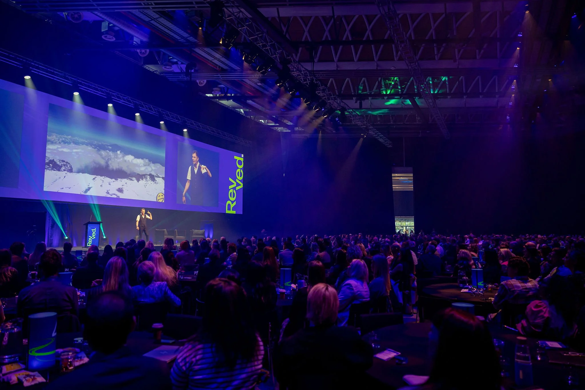 Revved conference audience seated in a darkened hall, listening to a speaker on stage with a large screen behind him showing mountain scenery and a person making a gesture. Stage is lit with blue and purple lights.