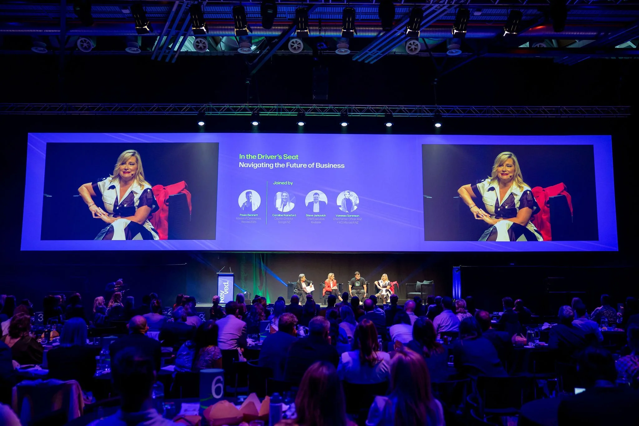 A large conference room filled with attendees sitting at tables, facing a stage with a big screen displaying a presentation titled 'In the Driver's Seat: Navigating the Future of Business' with images of four panelists and a panel discussion underway