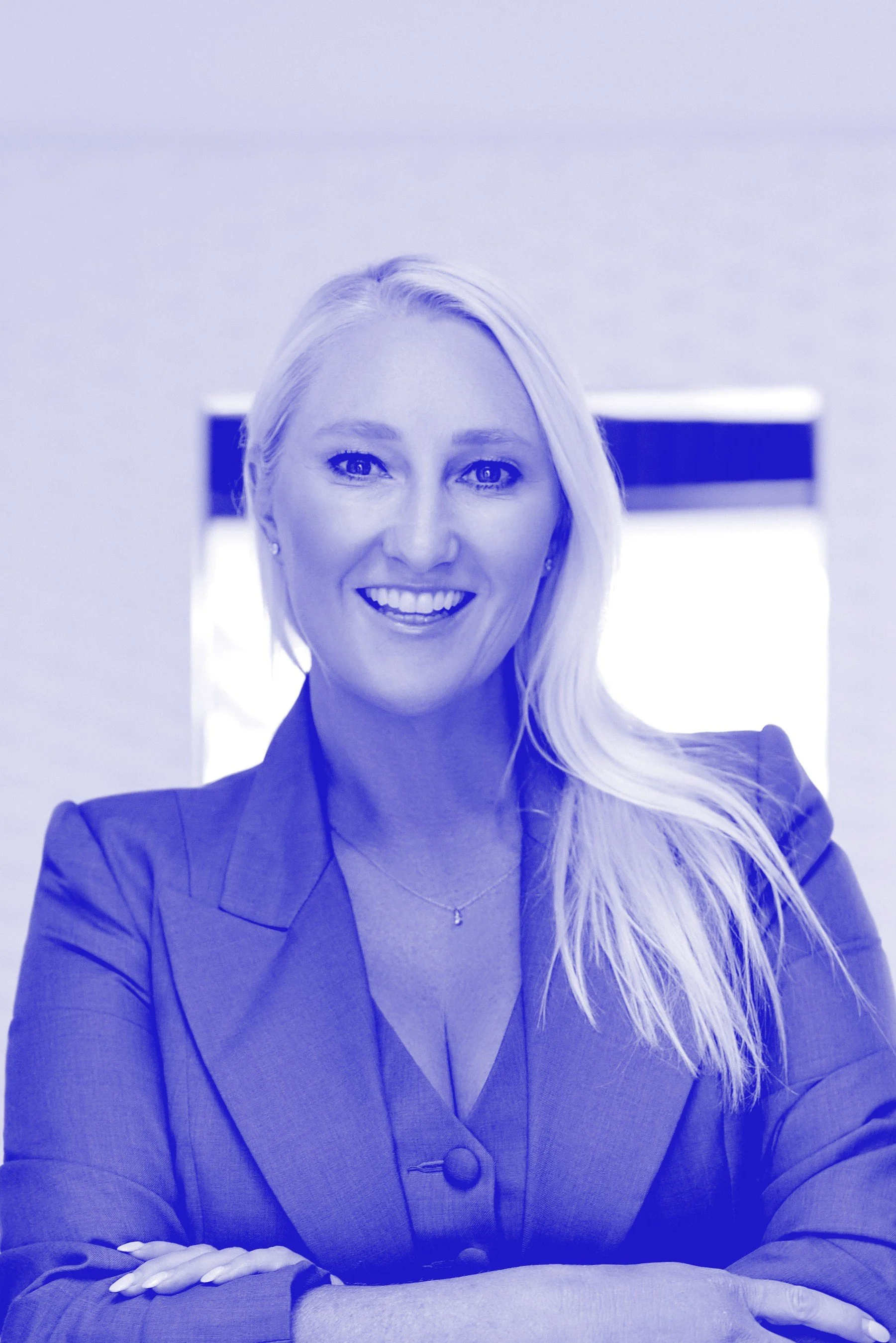 A professional woman with long blonde hair smiling in a business suit, sitting with her arms crossed.