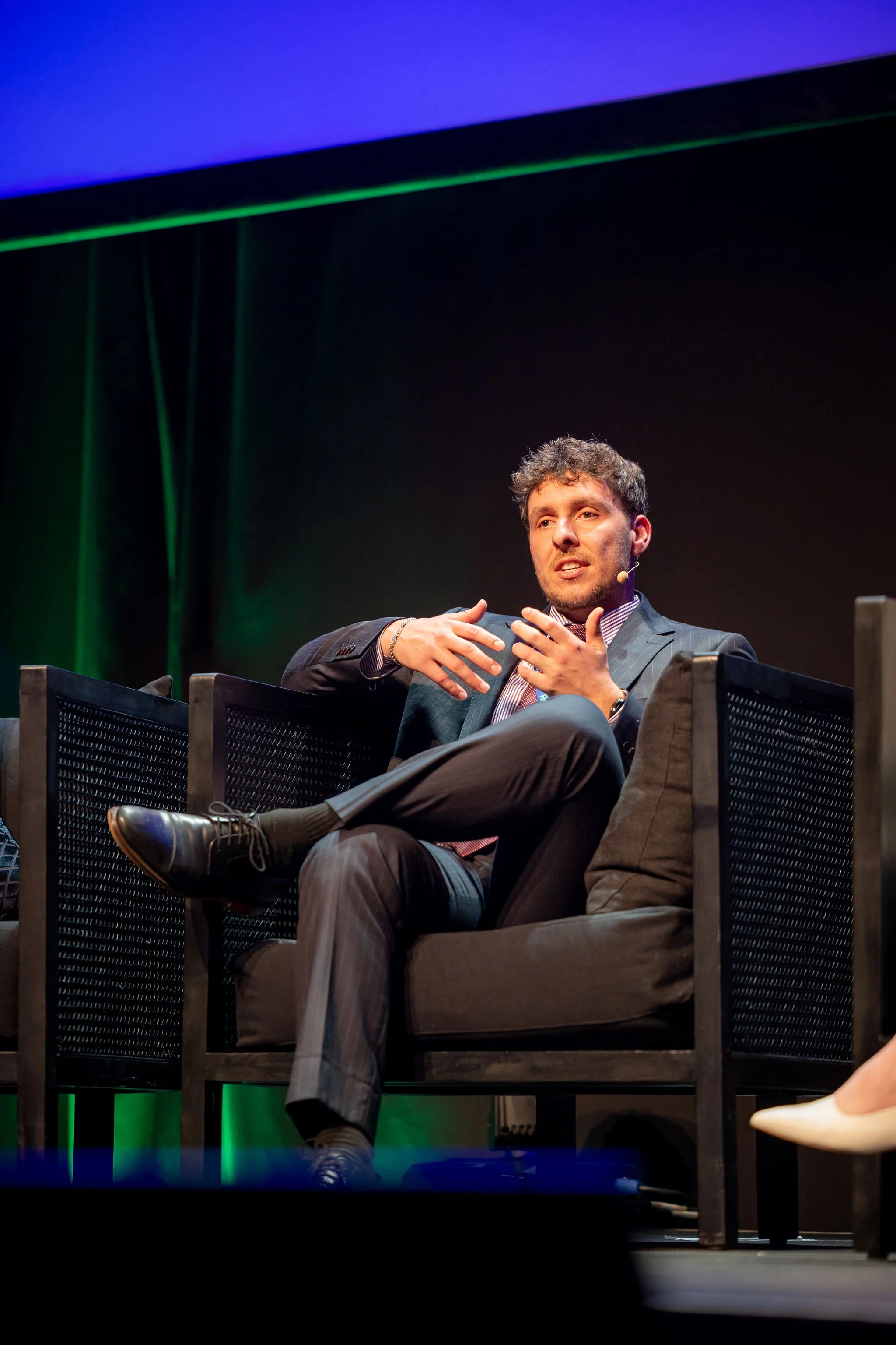 A man in a suit and striped shirt is sitting on a stage, speaking into a headset microphone, gesturing with his hands, with a dark background and stage lighting.