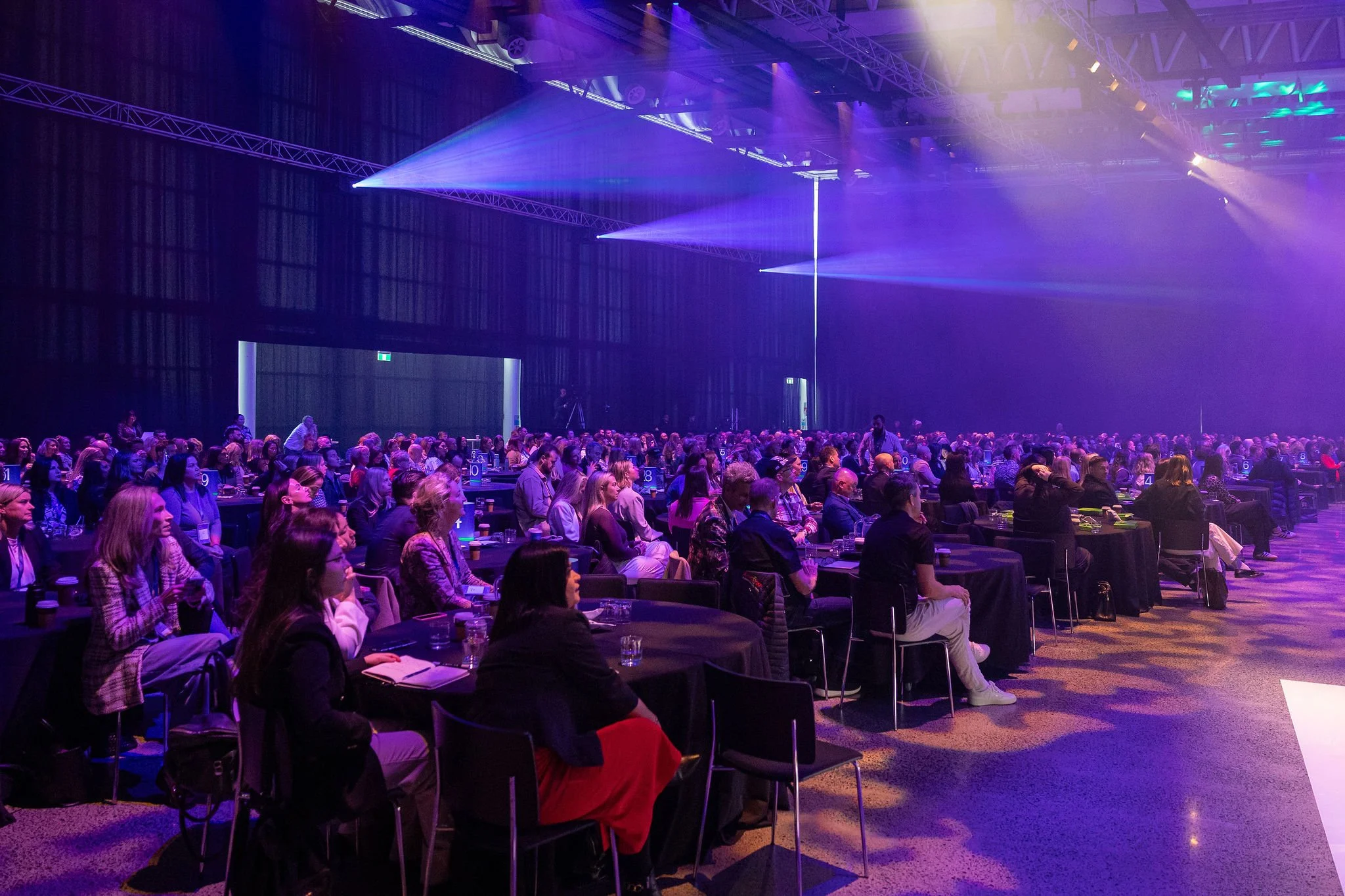 Revved audience seated at tables in a large conference hall with purple and green stage lighting.
