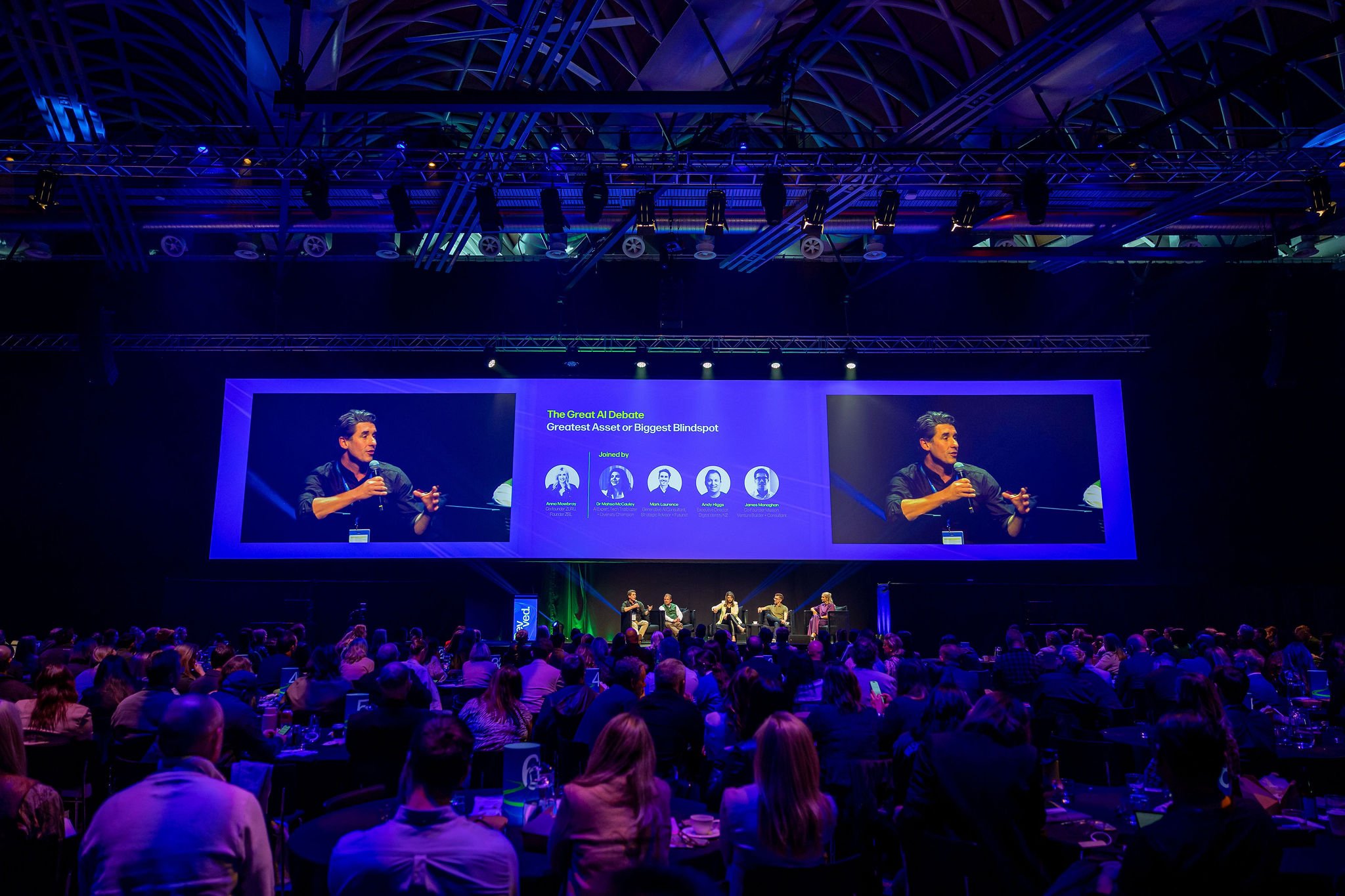 A conference or panel discussion at a large event with an audience seated at tables, a stage with a large screen showing a presentation titled 'The Great AI Debate,' and a panel of speakers seated on the stage.