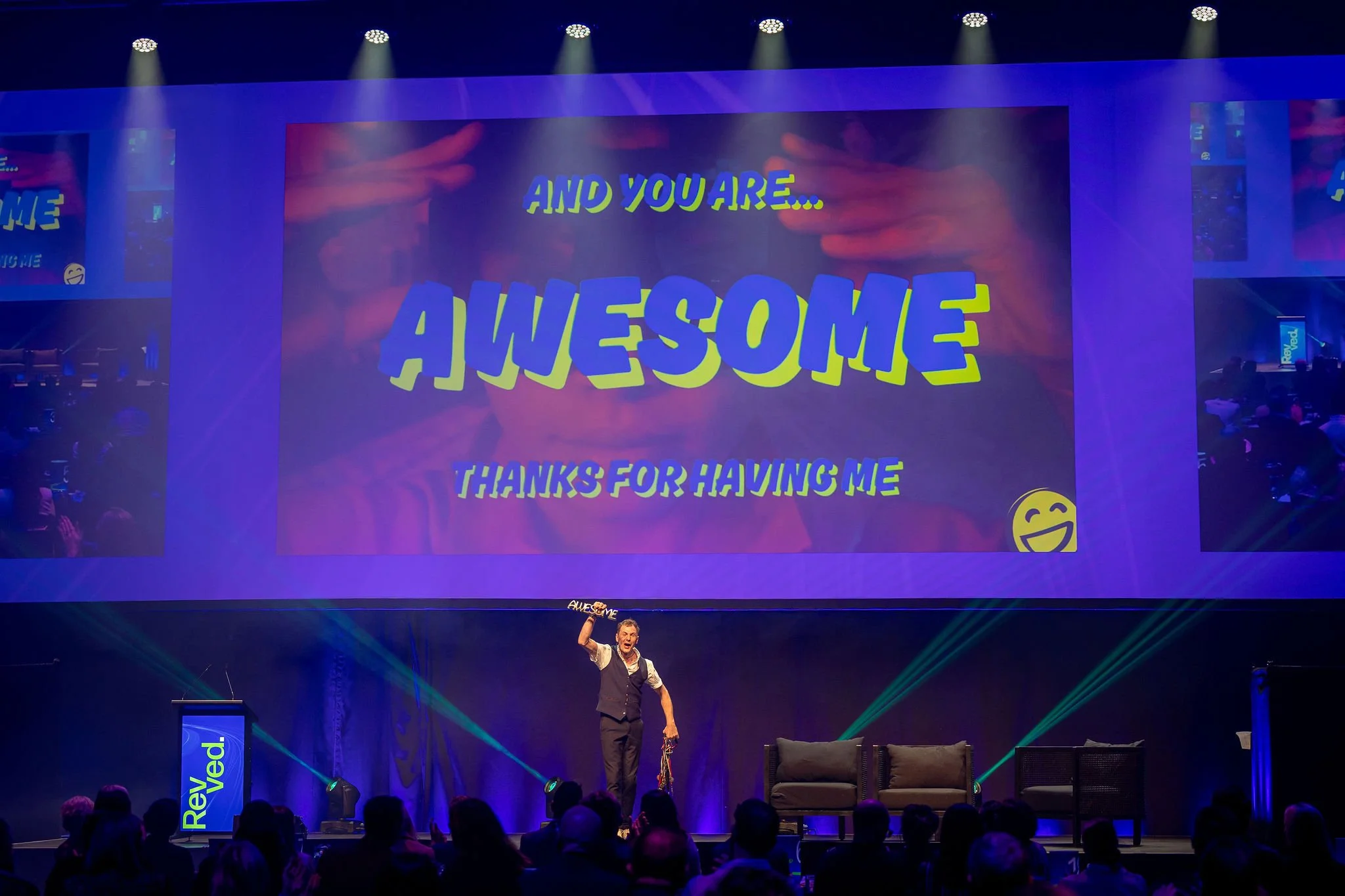 A speaker on stage at Revved with a large screen behind him displaying colorful text that says 'AND YOU ARE... AMAZING' and 'THANKS FOR HAVING ME' with a yellow smiley face emoji. 