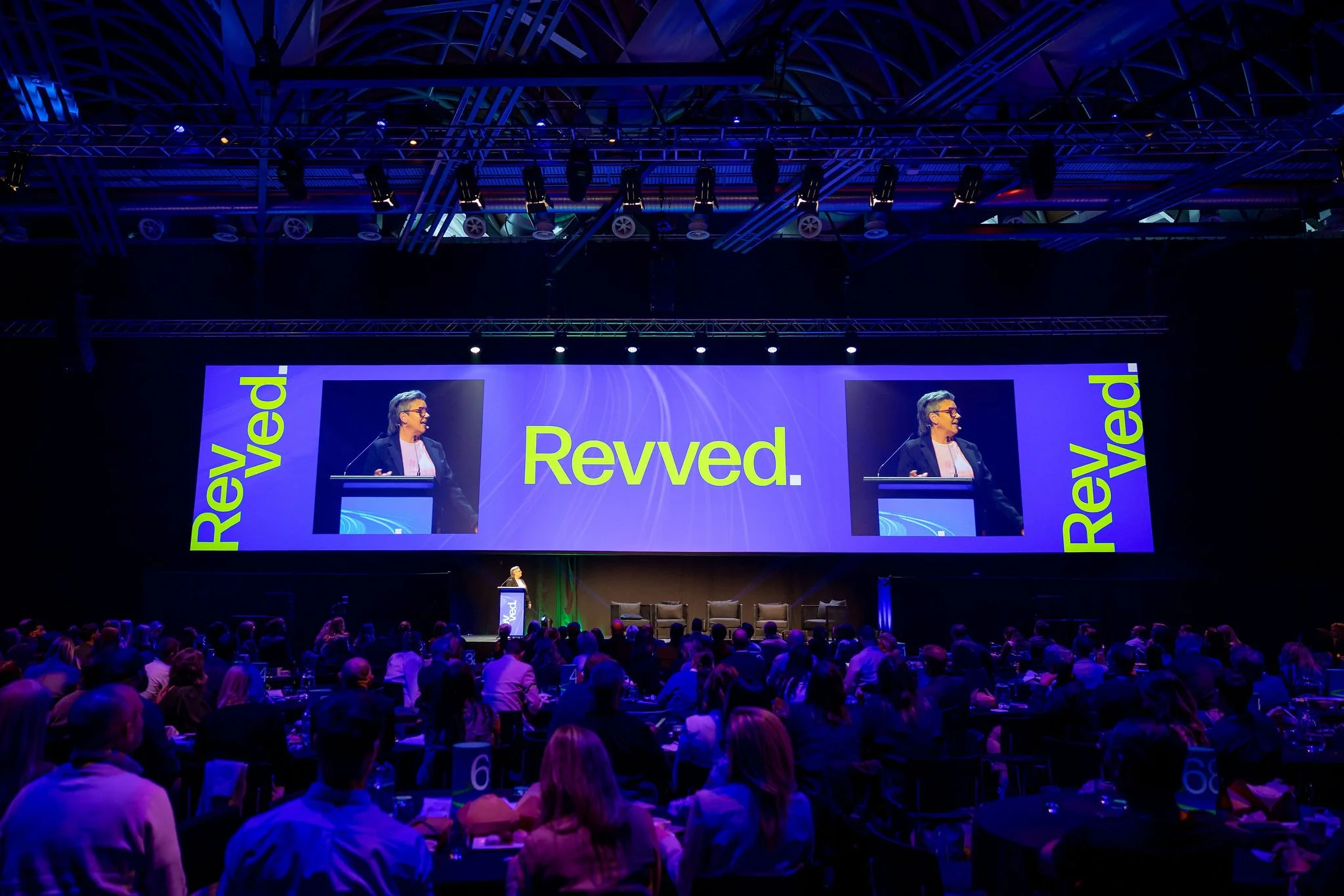 Large conference hall filled with attendees watching a speaker on stage. The stage has a gigantic screen displaying the word 'Revved.' and the speaker's image, with matching side visuals that say 'Revved.' in bright yellow and blue. The ceiling has n