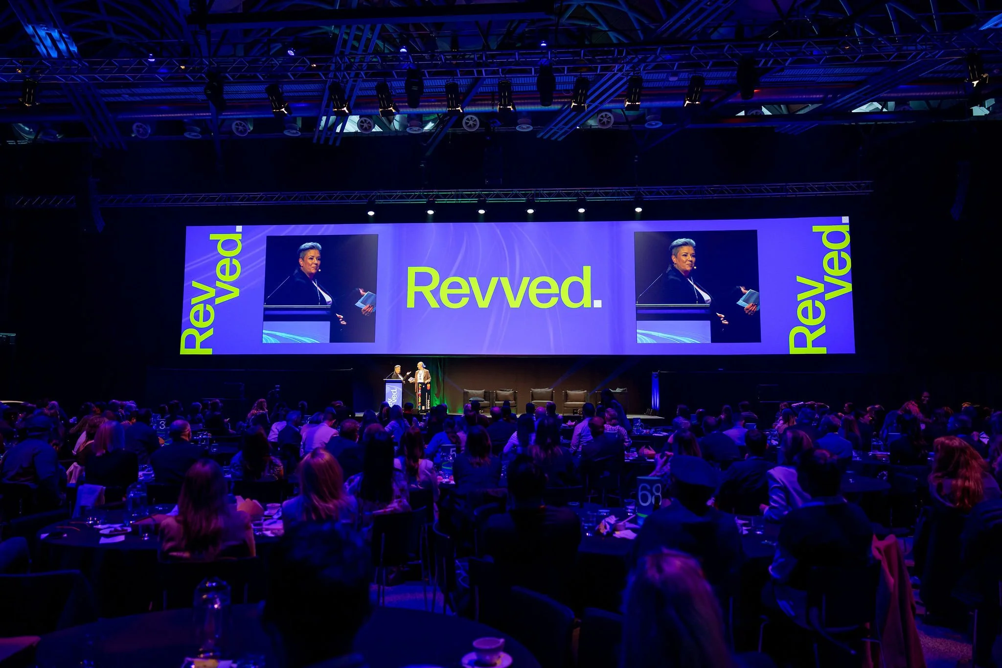 A large conference or event hall filled with attendees seated at tables, facing a stage with big screens displaying a woman speaking at a podium. The screens show the word 'Revved.' in large yellow letters on a purple background. The ceiling has an e