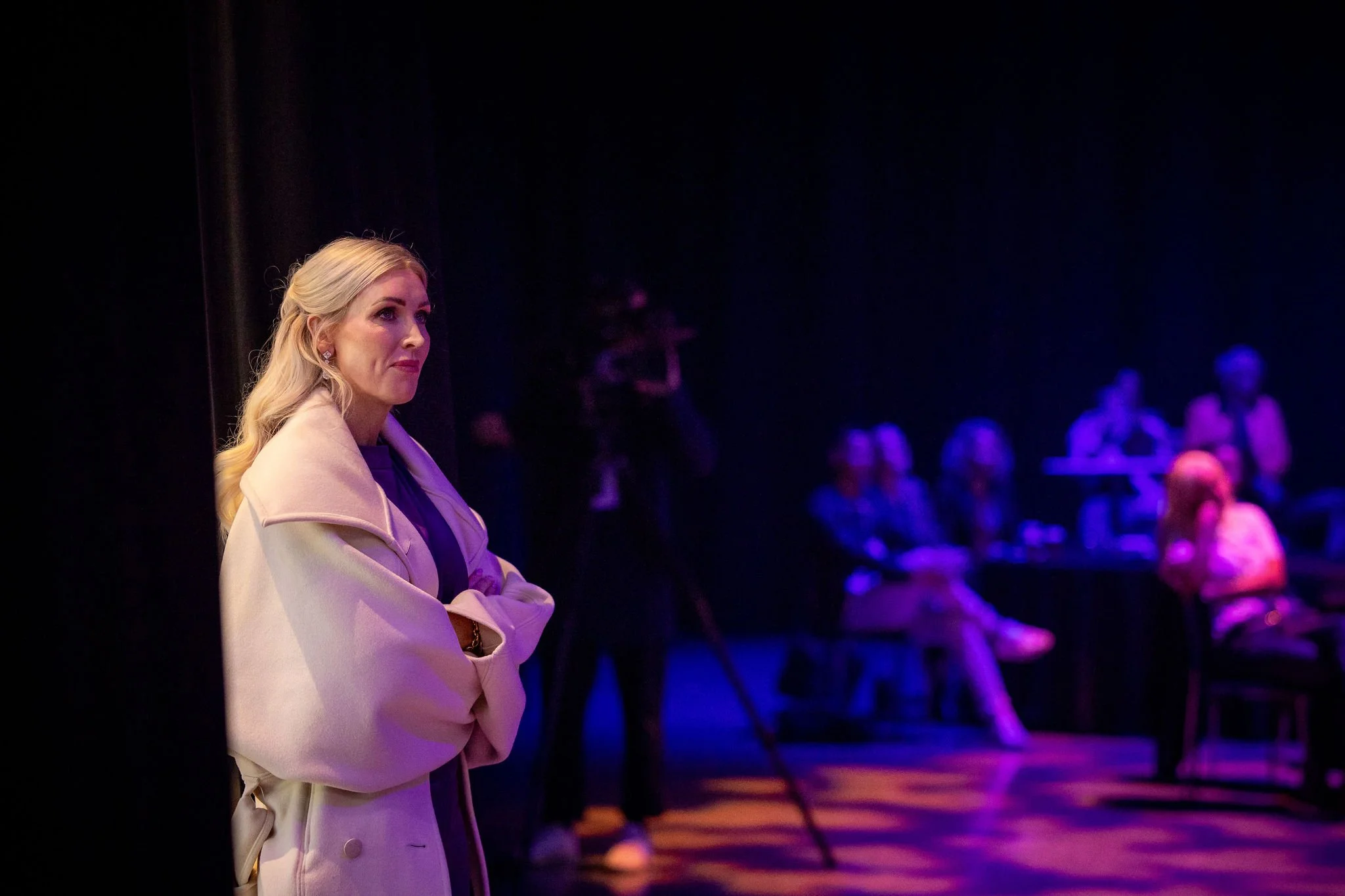 A woman with blonde hair and a light-colored blazer at Revved, with people sitting in the background in a room illuminated by purple and blue lights.