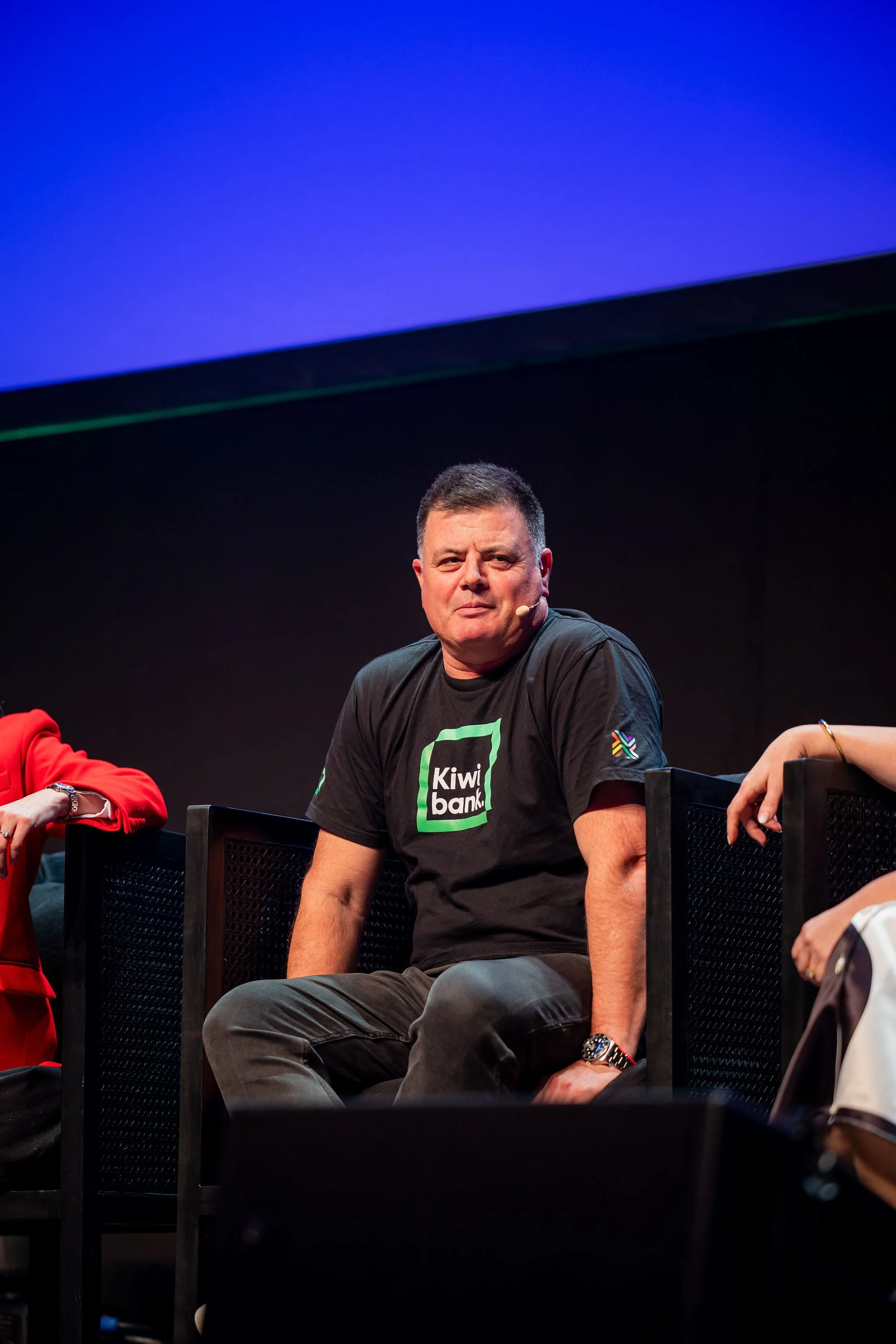 A man with short dark hair sitting on a stage in front of a dark background. He is wearing a black t-shirt with a logo that says 'KiwiBank' and has a rainbow-colored logo on the sleeve. He is looking at the camera and wearing a watch on his left wris