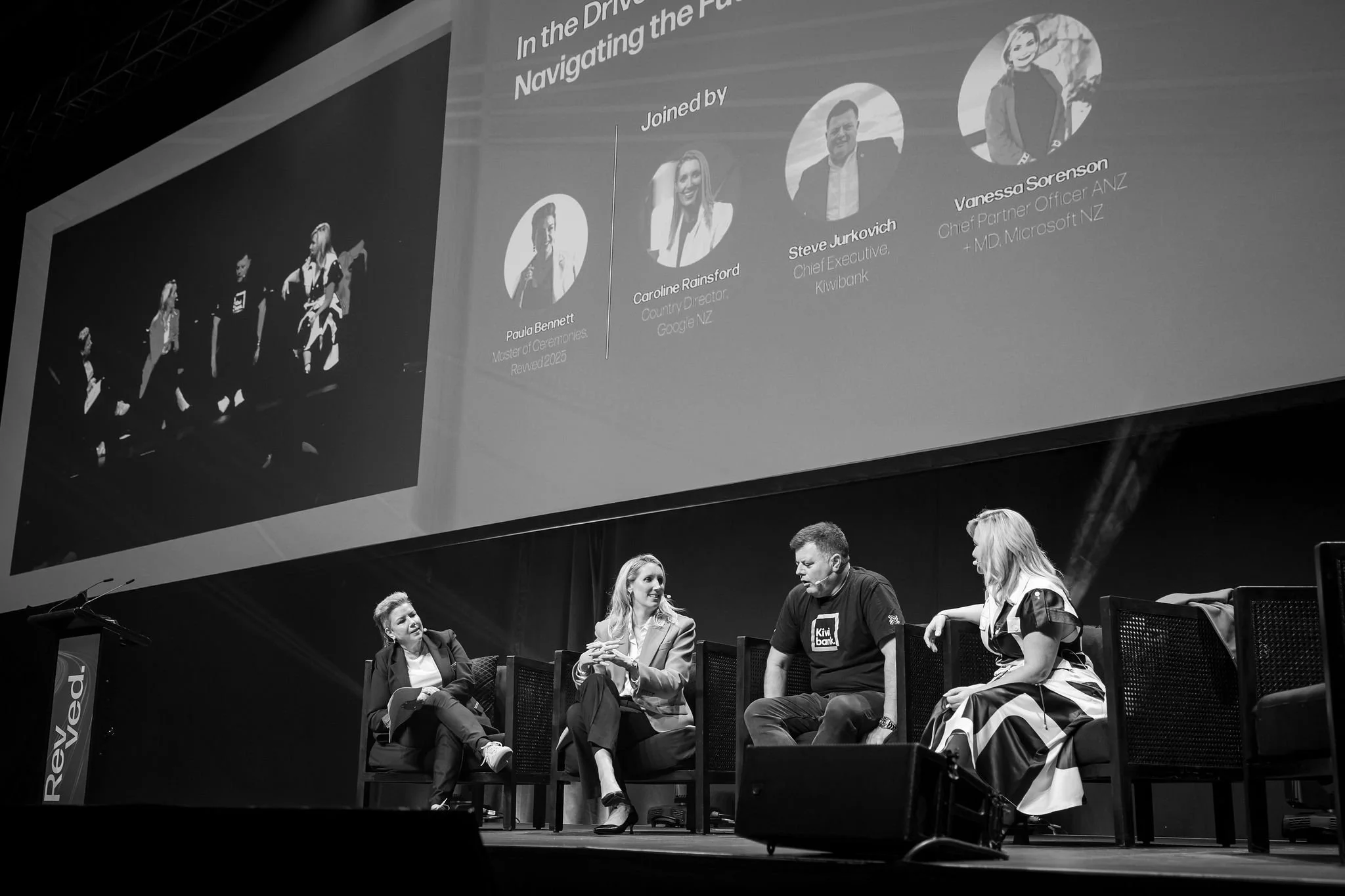 Several women and men seated on stage in discussion, with a large presentation screen overhead showing images and names of speakers at a conference or panel event.