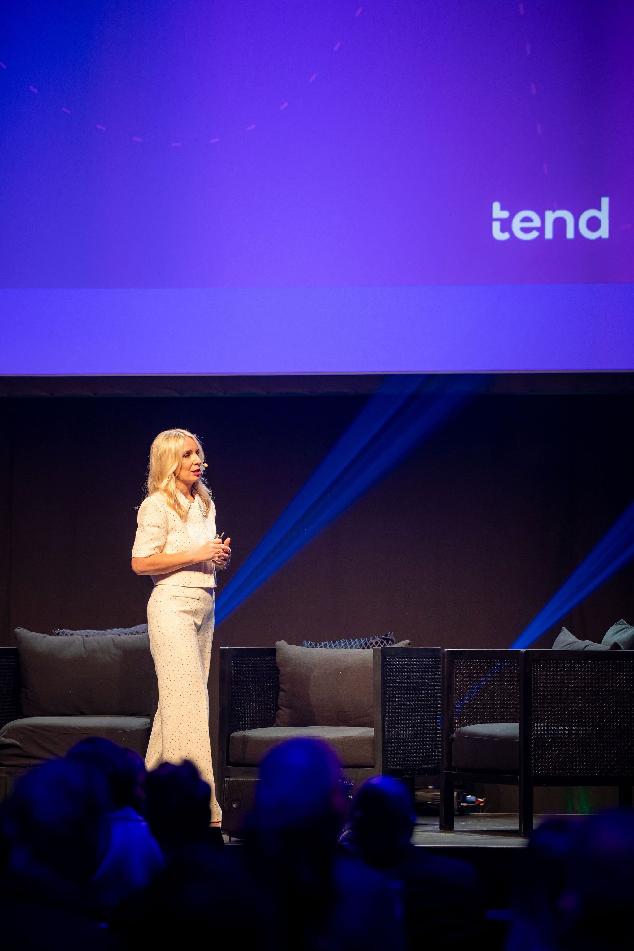 A woman with long blonde hair wearing a white polka dot suit is giving a presentation on stage. The background features a large screen with the partial word 'tend' and a dotted line circle design in purple hues. The stage has black furniture and audi