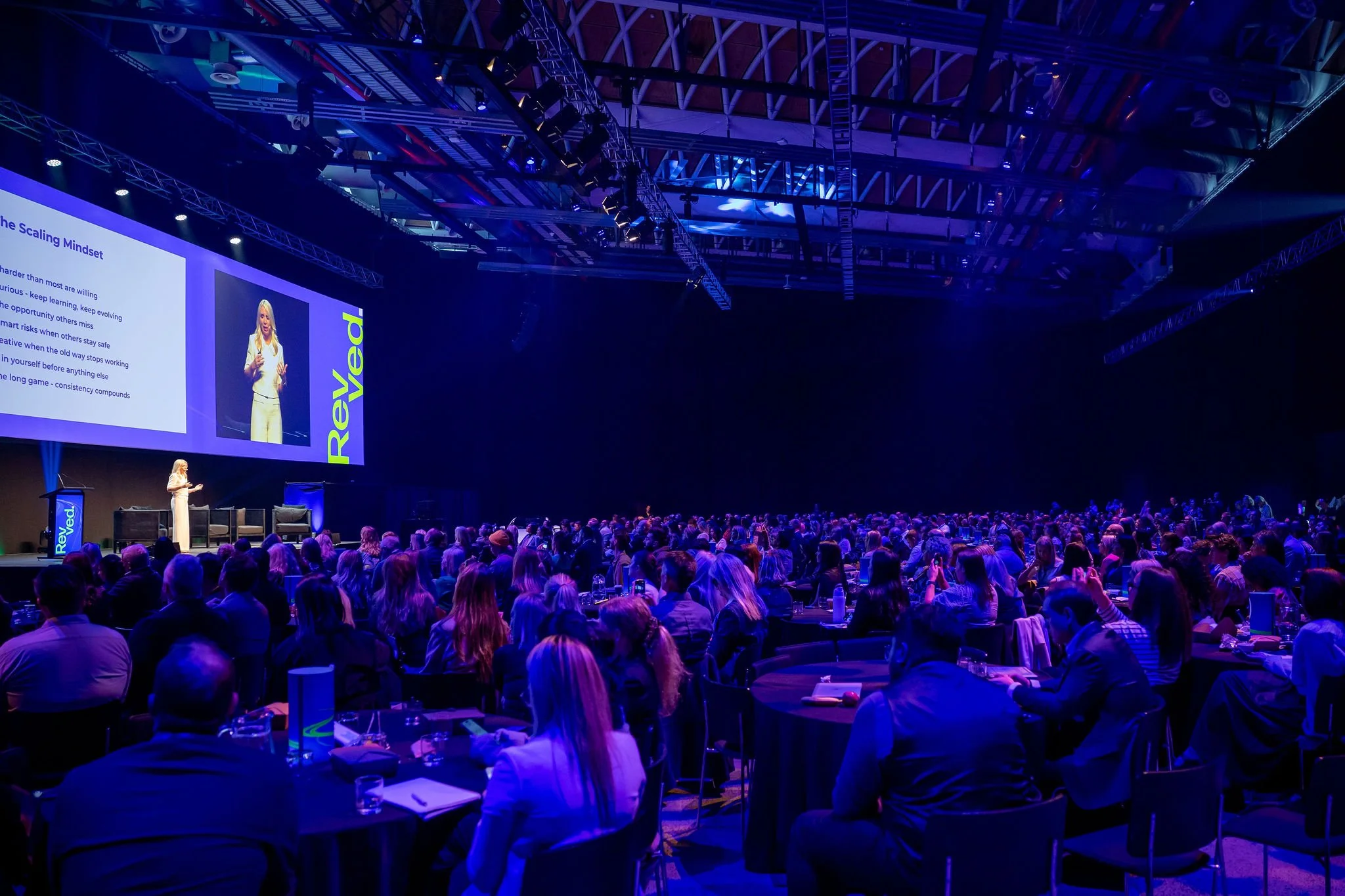 Large conference hall filled with seated attendees listening to a woman speaking on stage. The stage features large screens displaying presentation slides and the word 'Reyved' on the side. The ceiling is decorated with metal trusses and stage lighti