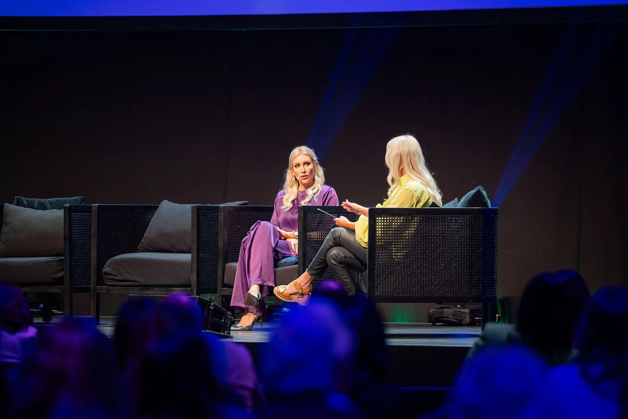Two women seated on stage, engaged in conversation, with many audience members watching in the foreground. One woman wears purple clothing and the other wears a yellow top and black pants.