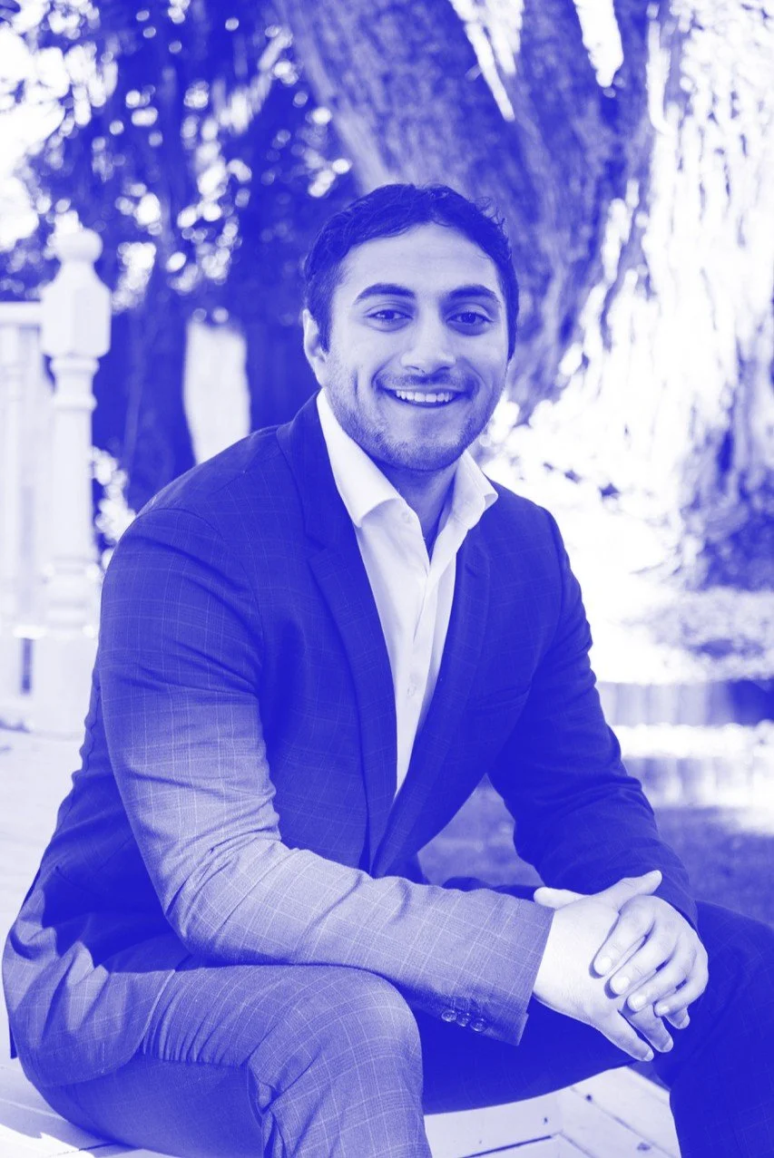 A smiling young man in a suit sitting outdoors with trees and a white railing in the background.