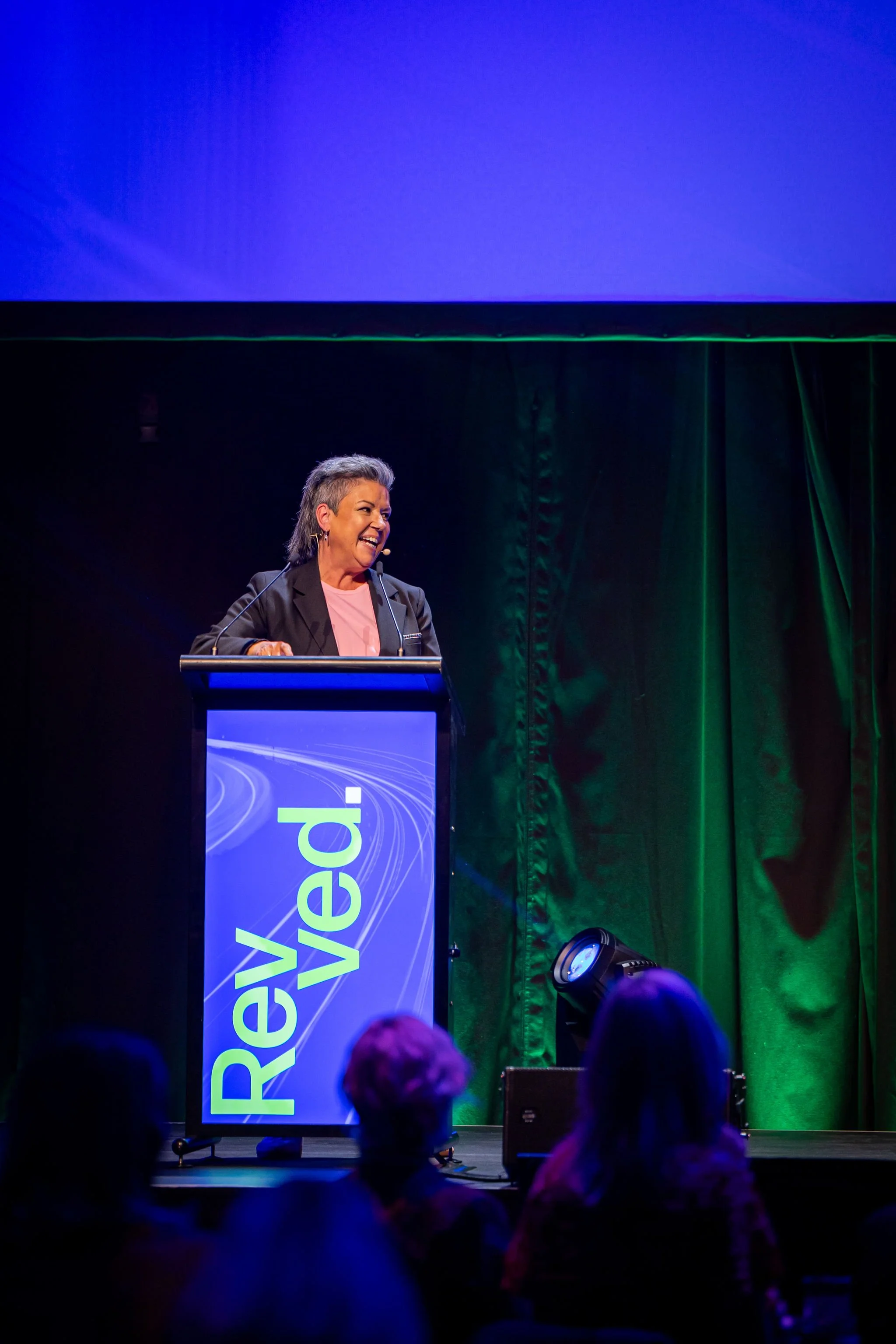 A woman with short, gray hair speaking at a conference, standing behind a podium with a large screen displaying the words "Revent" in blue and yellow background, with a dark green curtain in the background.