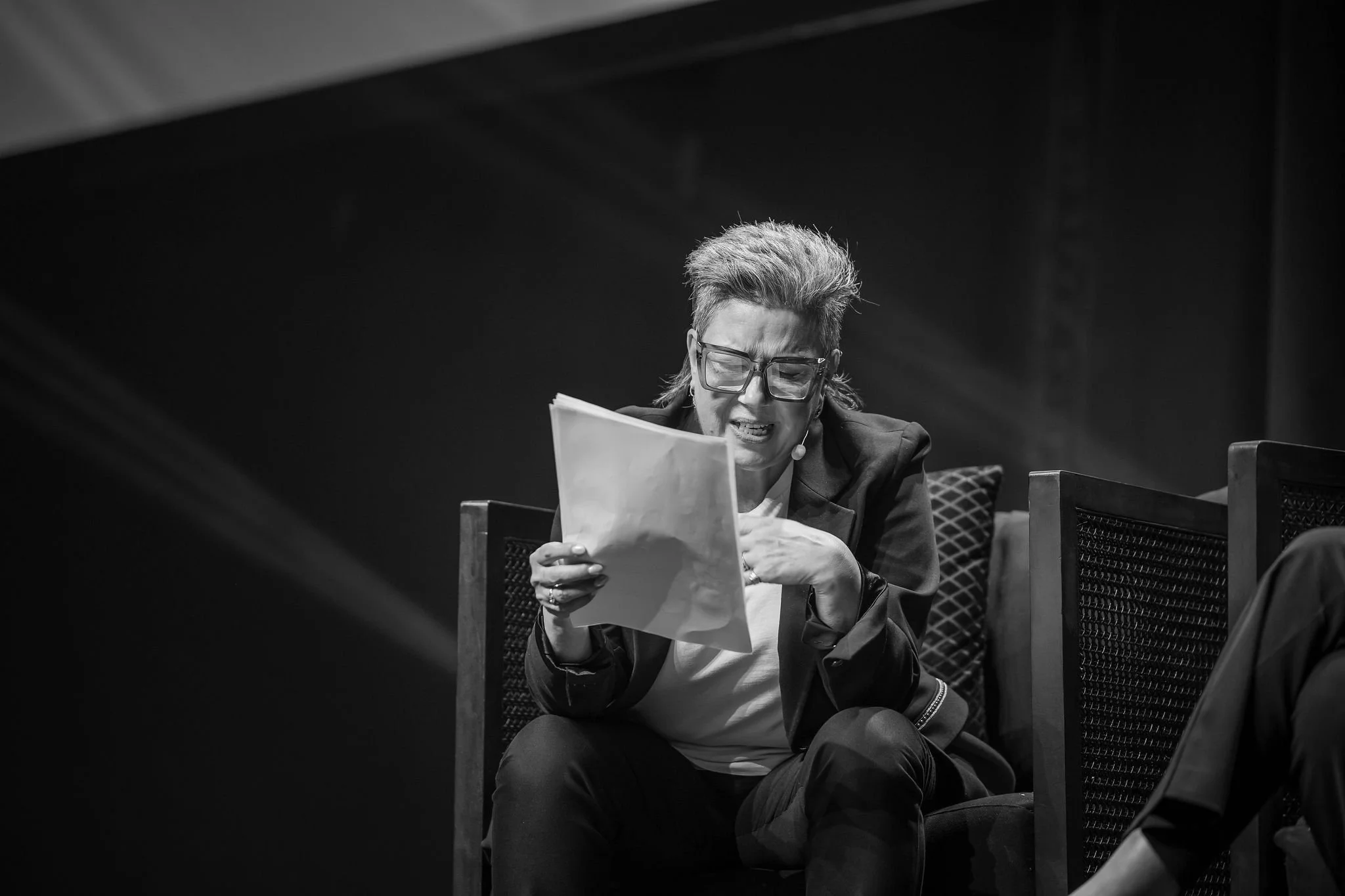 Woman with glasses and short hair, wearing a headset, appears to be frustrated or upset, reading a document while sitting on a chair in a dimly lit room.