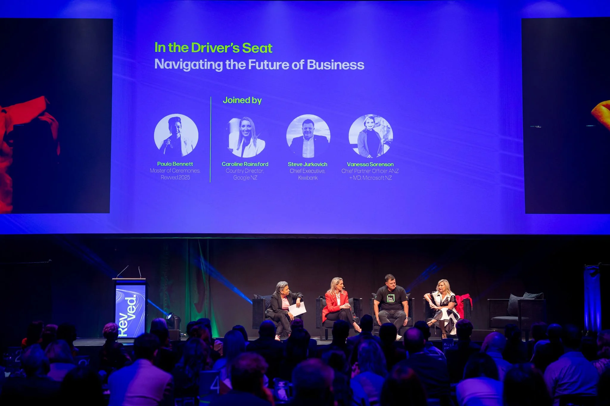 Panel discussion at a conference with five seated speakers on stage and a large screen behind them displaying the panel titles and photos of four speakers.