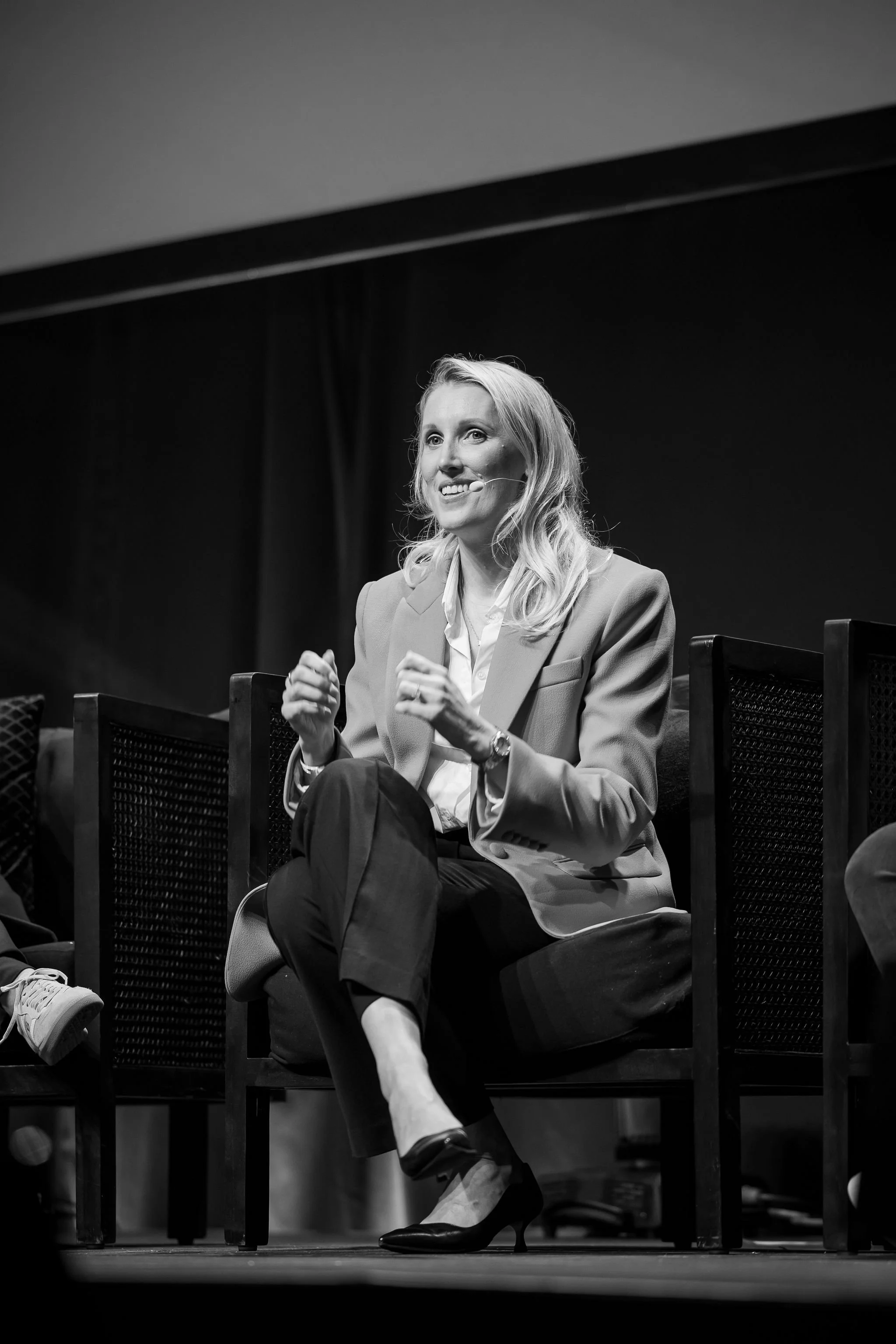 Black and white photo of a woman sitting on a chair on stage, speaking into a headset microphone. She is wearing a suit and high heels, with her legs crossed, and appears to be engaged in a panel discussion or presentation.