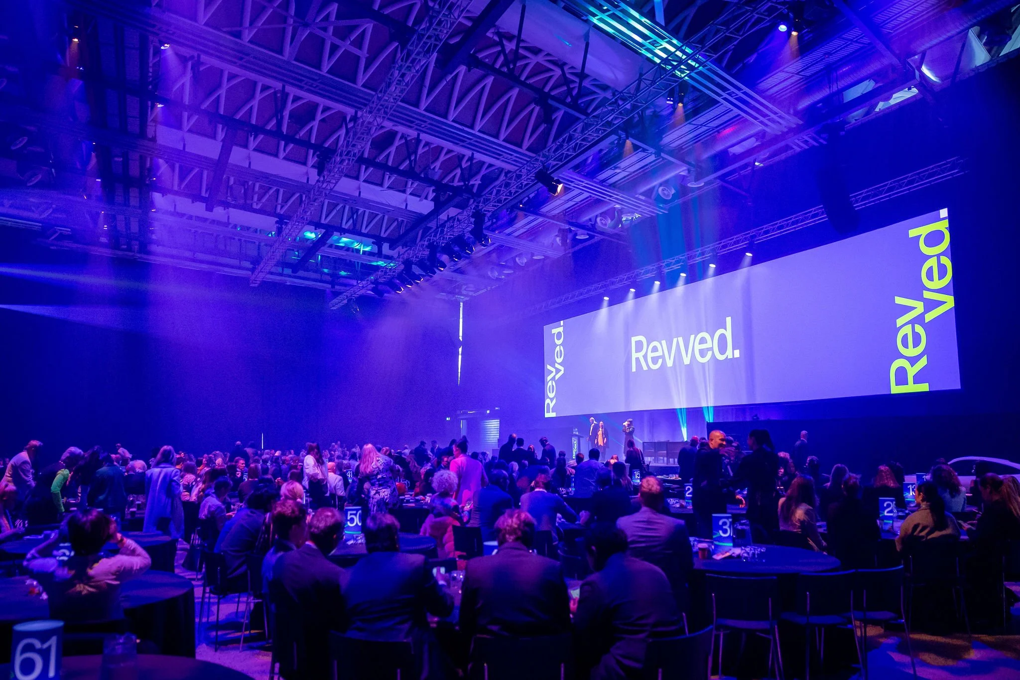 Revved conference auditorium filled with attendees seated at tables, facing a stage with large screens displaying the text "Revved." and other words, illuminated with purple and blue lighting.