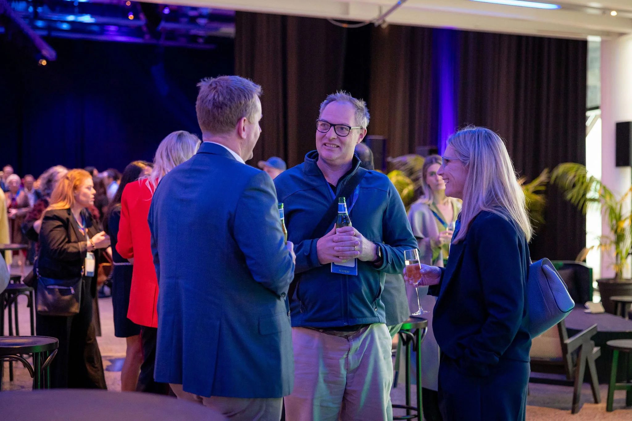 Three people are engaged in conversation at a social event. The man in the middle is holding a beer and is smiling, wearing glasses and a gray jacket. The man on the left, dressed in a suit, has his back to the camera, and the woman on the right is h
