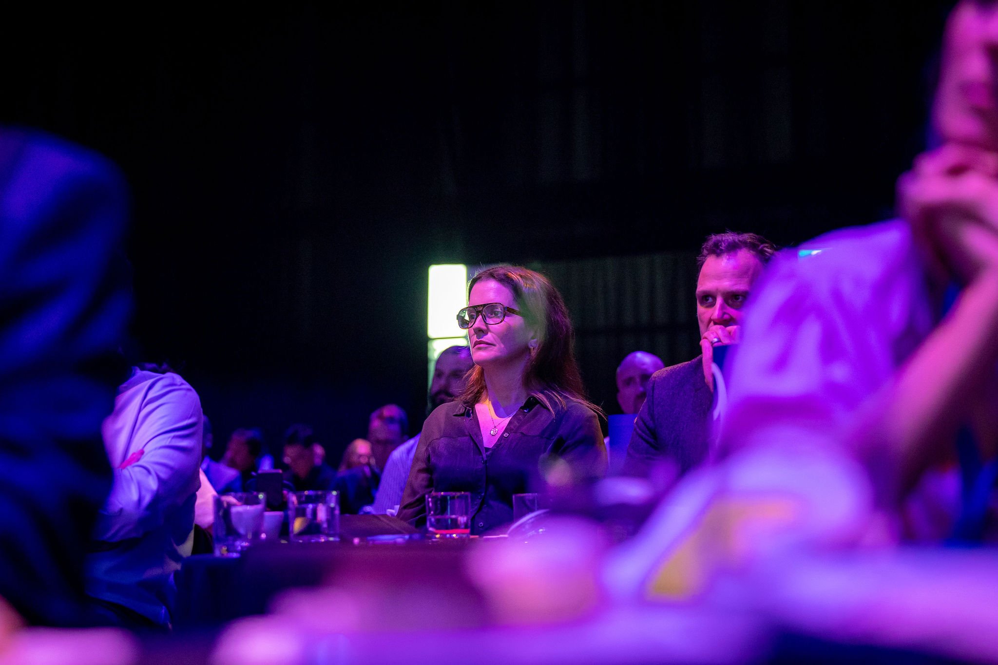 A woman with glasses and reddish-brown hair is seated at a table in a dark, colorful event setting. She appears to be listening attentively, surrounded by other people. There are drinks on the table, and the lighting is dim with neon purple and green