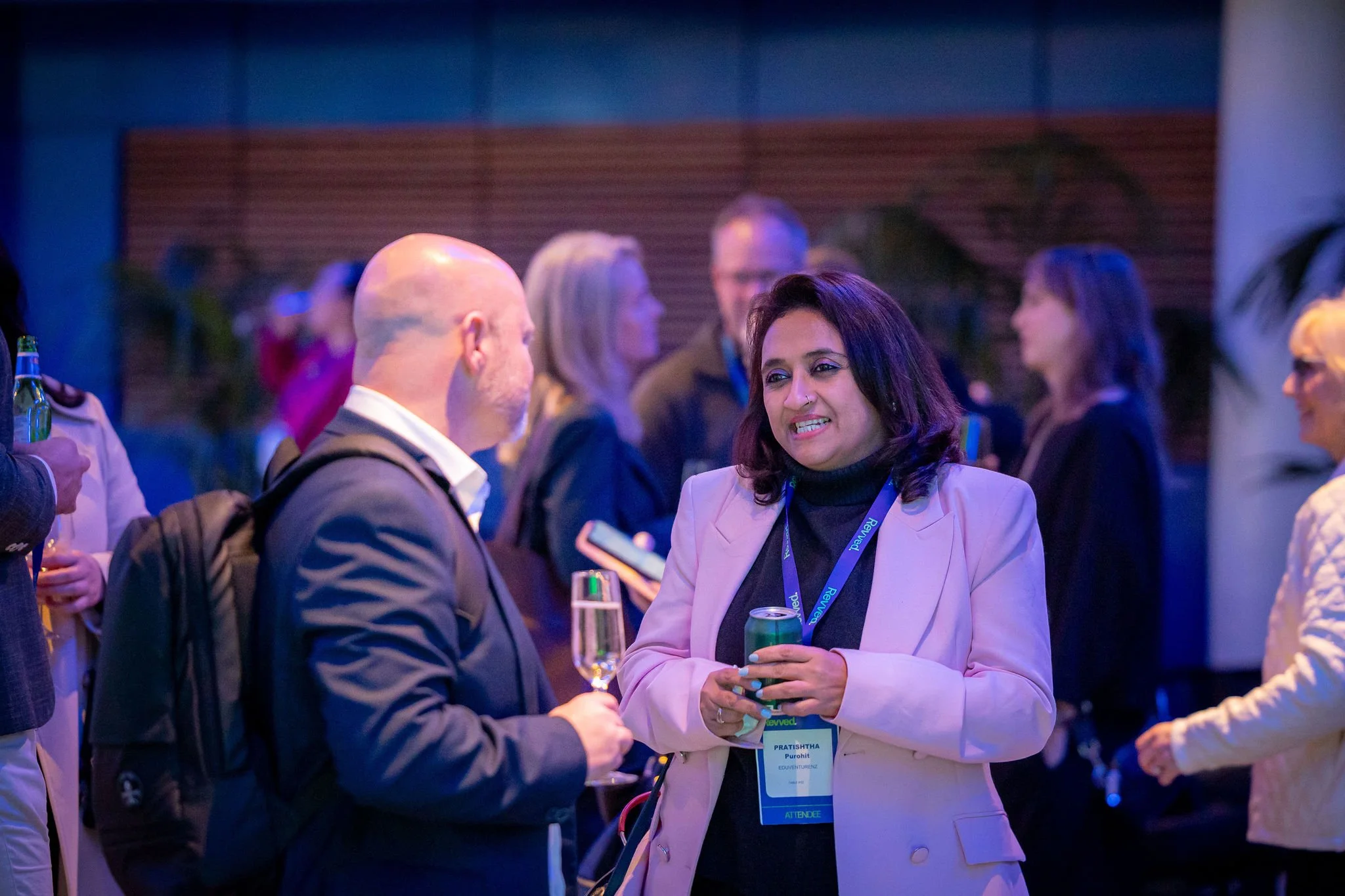 A woman in a white blazer and black turtleneck is talking with a man at a conference or networking event. The woman has dark hair, wears a conference badge, and is holding a green drink. The man is bald with a beard, wearing a dark suit, and carrying