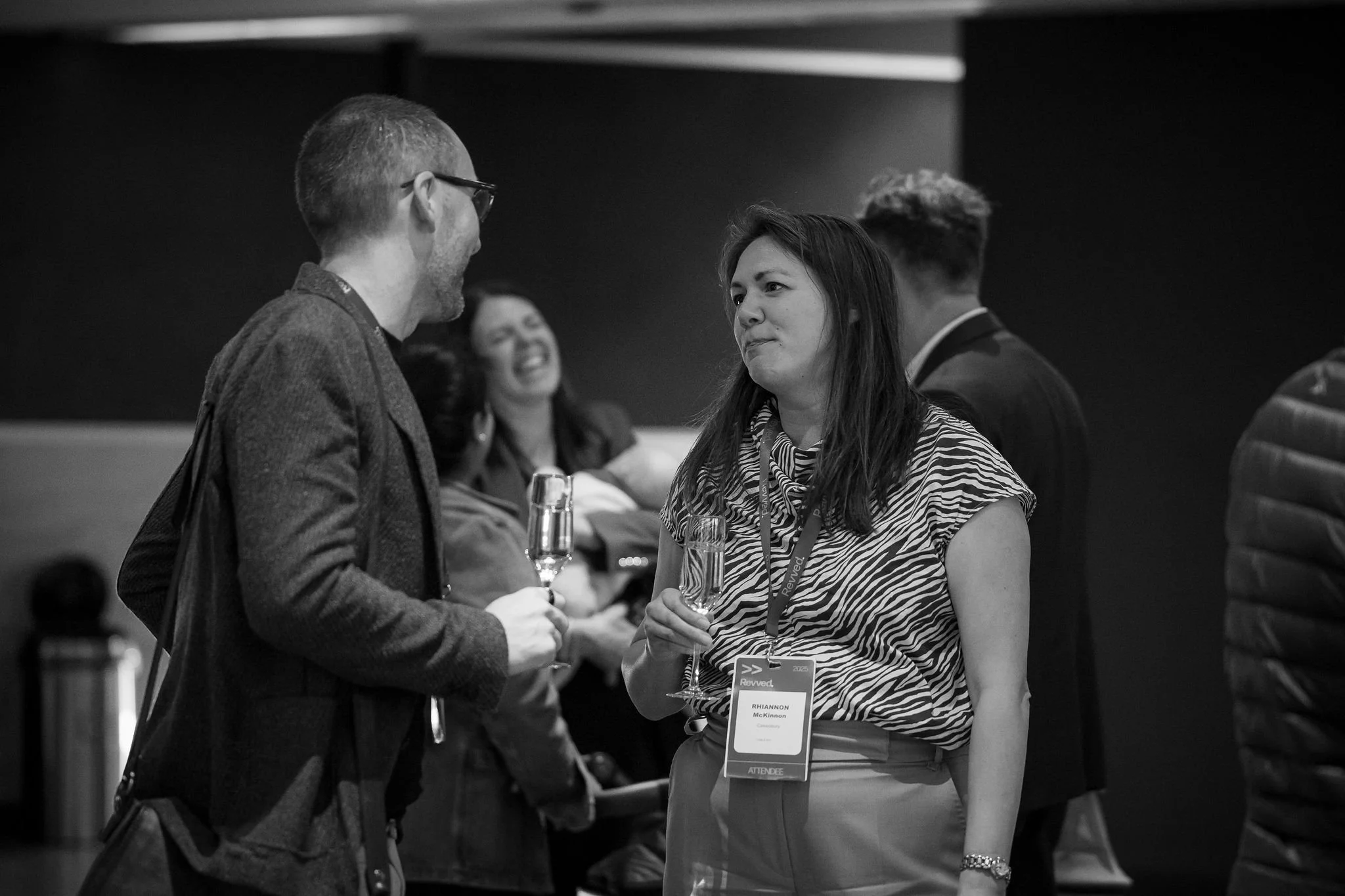 Two men and a woman engaged in conversation at a professional event. The woman, wearing a name badge and holding a glass of wine, is smiling and facing the man on the left, who is also holding a glass of wine. Another person can be seen in the backgr